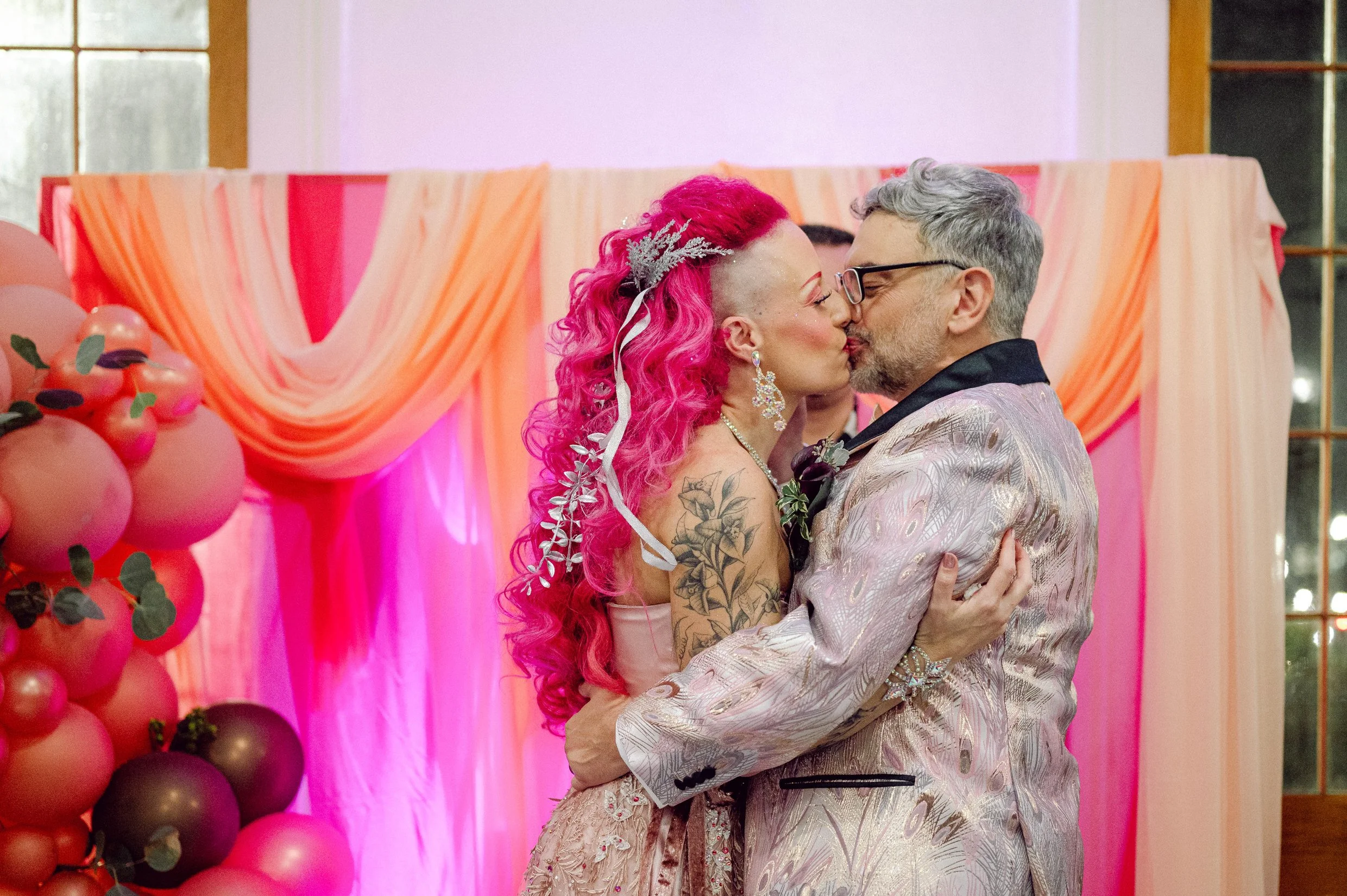 A bride and groom kisses their first married kiss in front of pink and peach draped decorations, with balloons and greenery on the side.