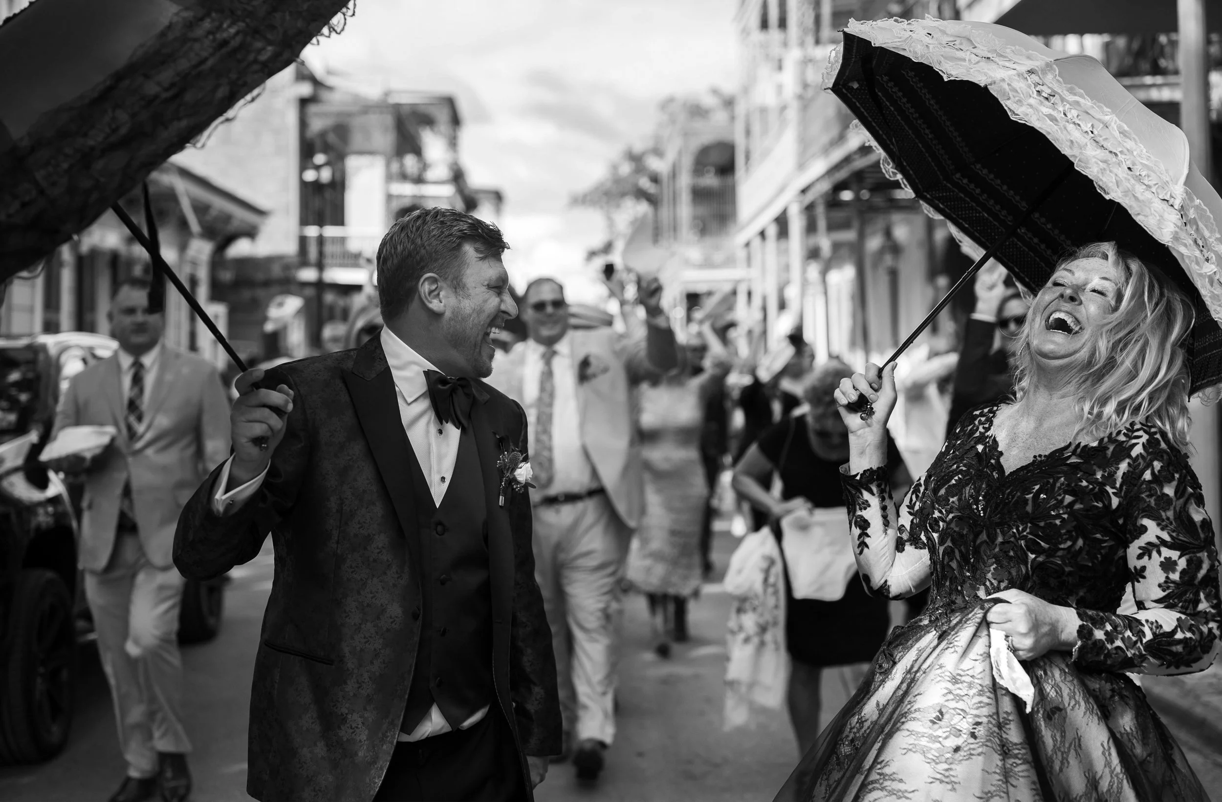 A bride and groom dancing in their wedding second line, with a man in a tuxedo smiling and a woman in a black wedding dress laughing under a parasol.