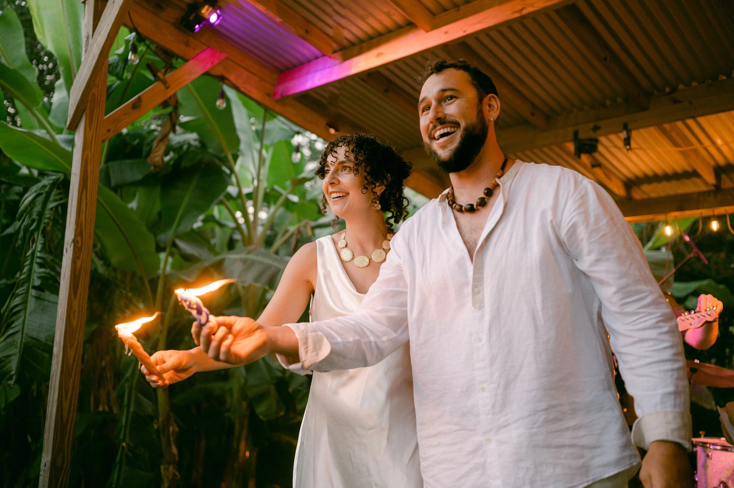 A bride and groom enjoying a casual celebration outdoors, holding sparklers, with tropical plants and string lights in the background.