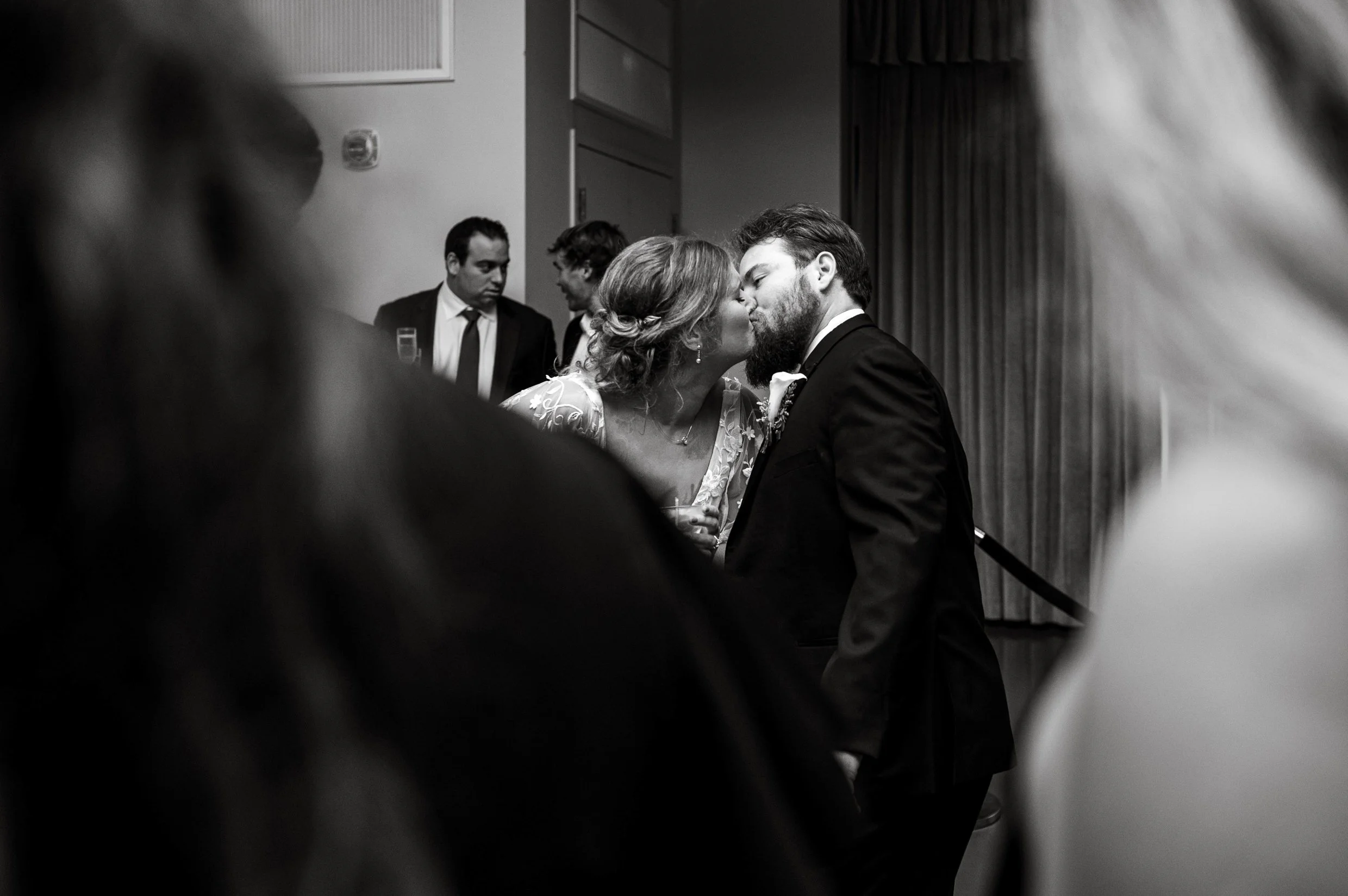 A black and white photograph of a couple sharing a kiss at their wedding , and the image framed by blurred figures in the foreground.