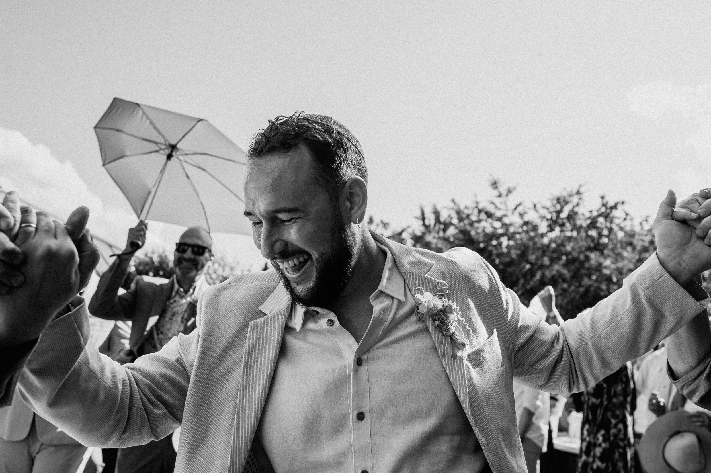 A groom smiling and dancing at his wedding surrounded by others, with trees and a partly cloudy sky in the background.