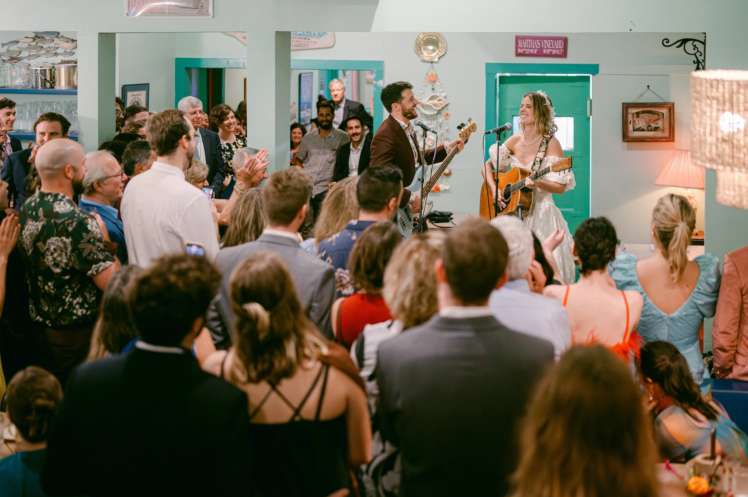 A bride and groom performing a song with guitars at their wedding reception, surrounded by joyful guests clapping and watching.
