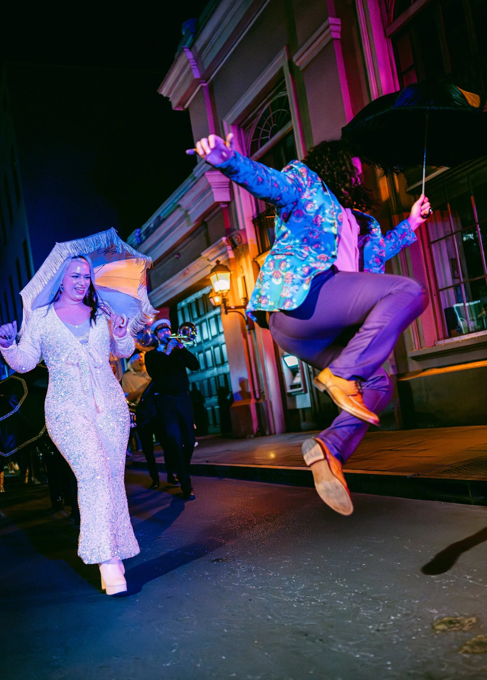 A groom mid-air jumping in front of a bride in a sparkly jumpsuit, at night illuminated by colorful lights in New Orleans.
