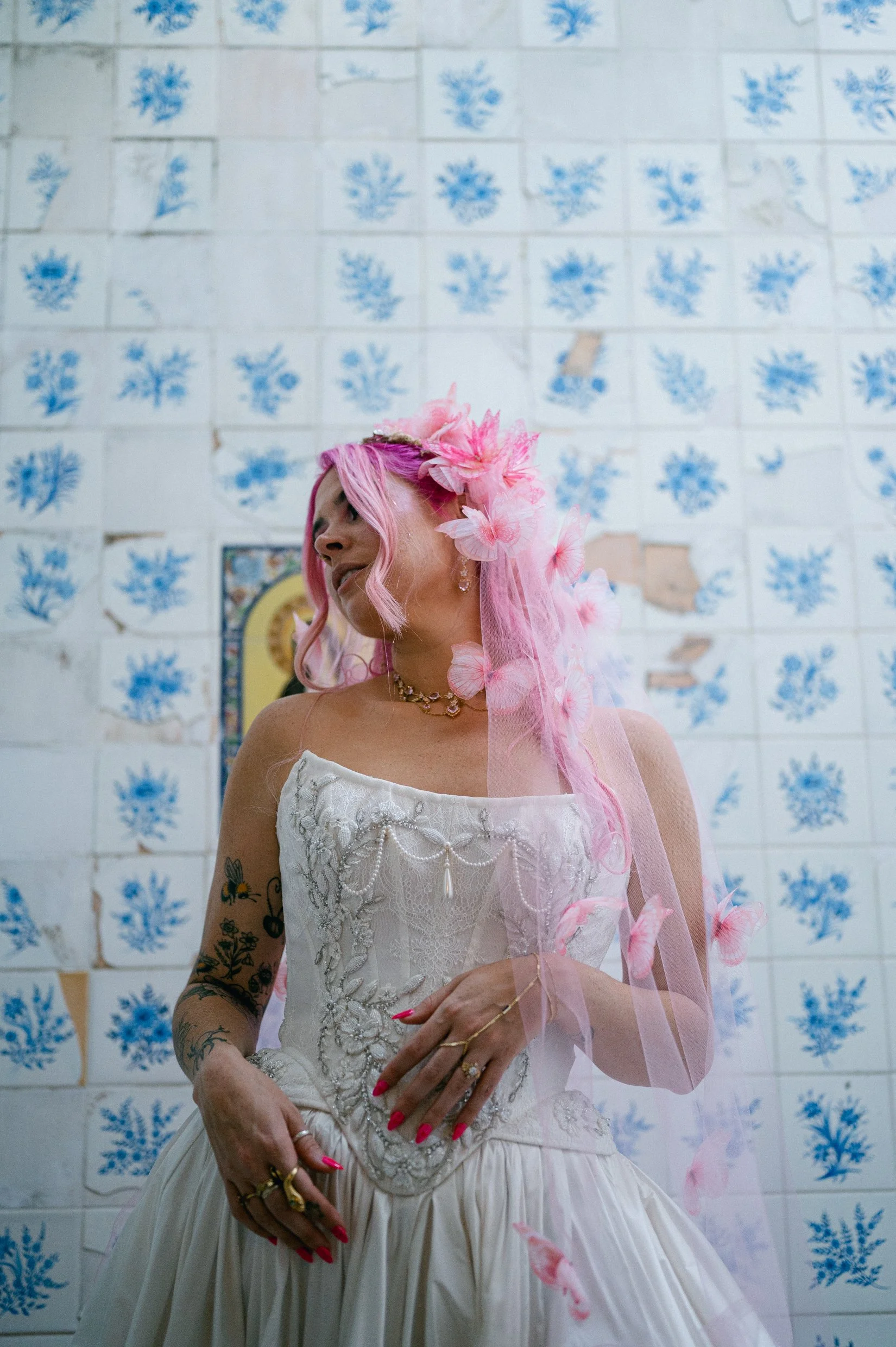 A woman with pink hair wearing a wedding dress and pink floral accessories stands in front of a wall with blue floral tiles.