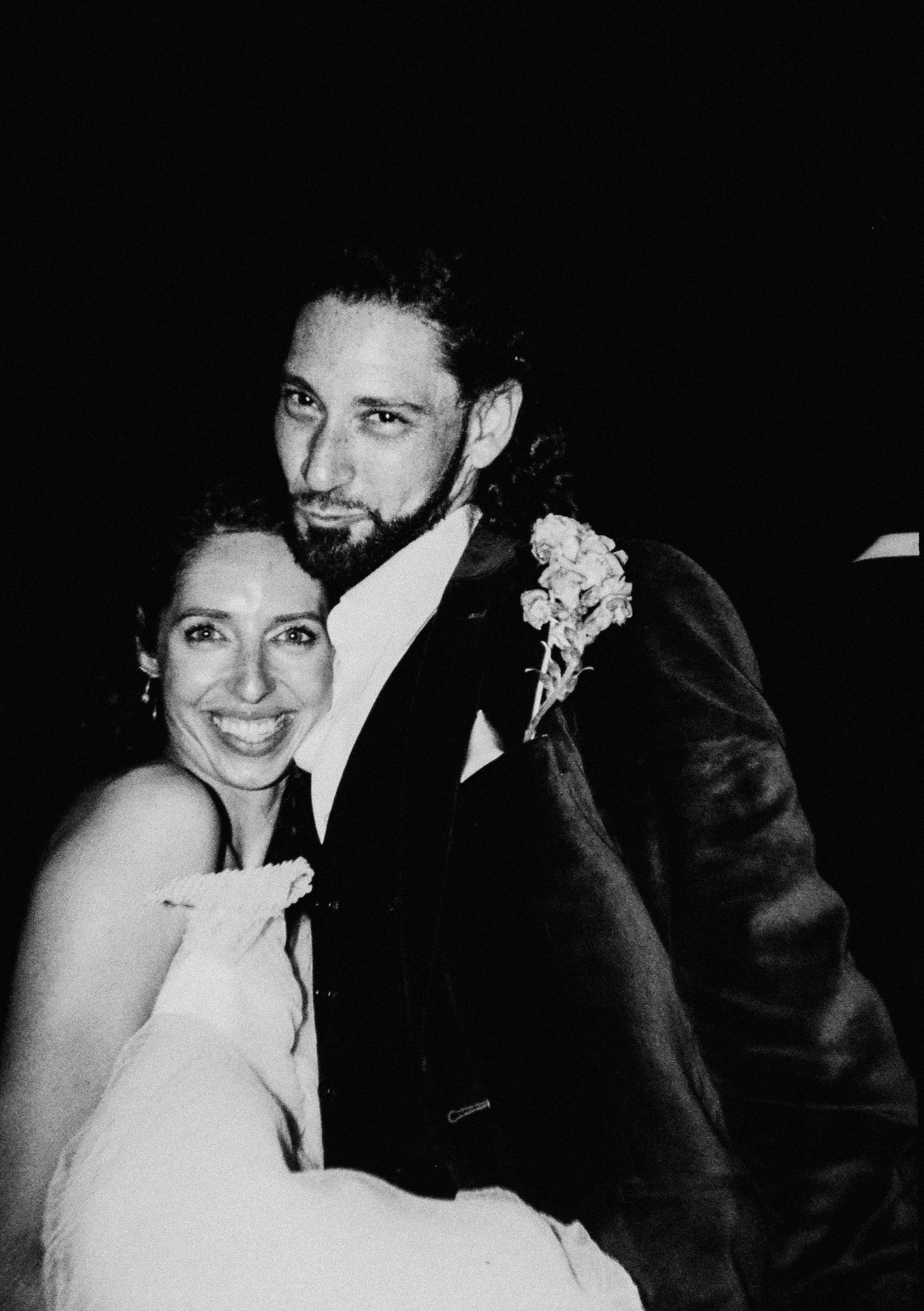 Black and white film photo of a couple dressed in wedding attire, smiling and embracing and looking directly at the camera.
