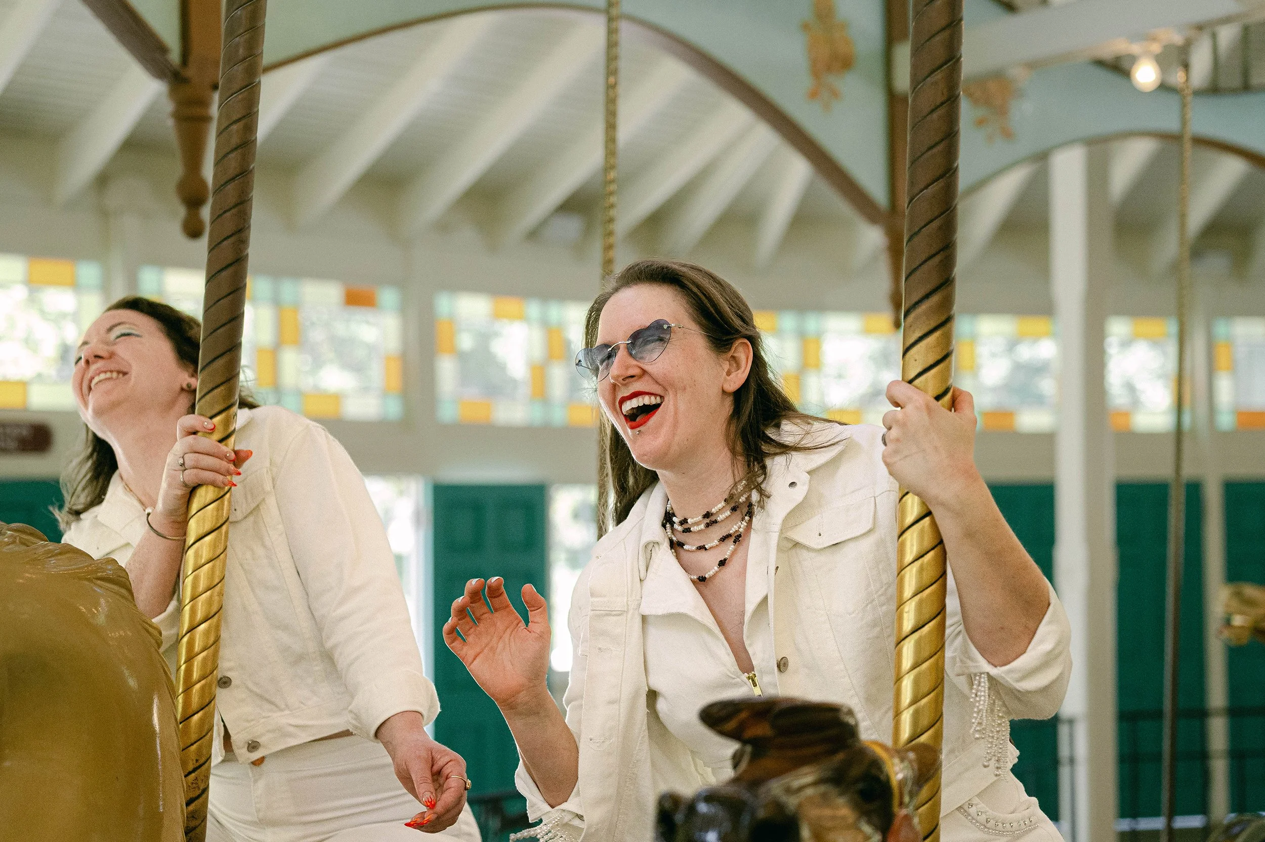 Lesbian newlyweds sitting on a carousel ride, smiling and laughing. 