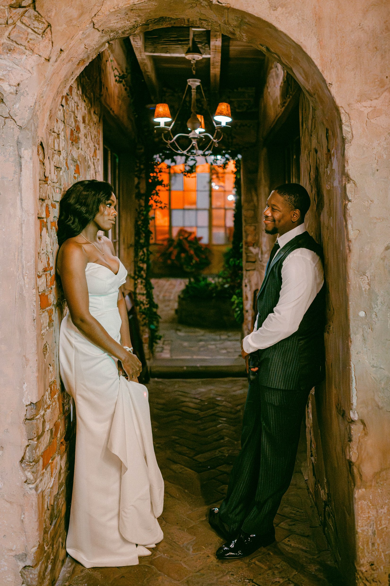 A black bride and groom standing in a rustic, brick archway hallway with dim lighting and a chandelier, facing each other.