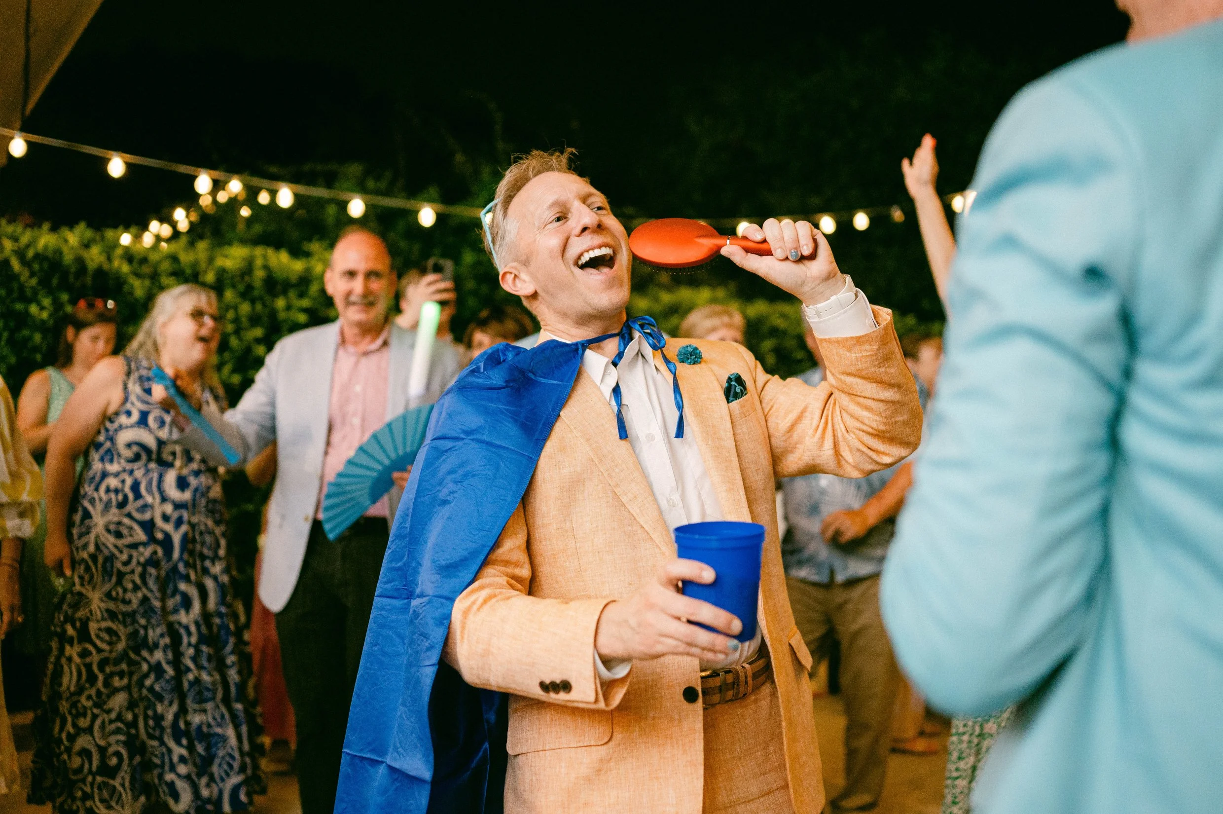 A groom at his wedding singing into a hairbrush and wearing a blue cape. Other guests are visible in the background, enjoying the evening with string lights overhead.