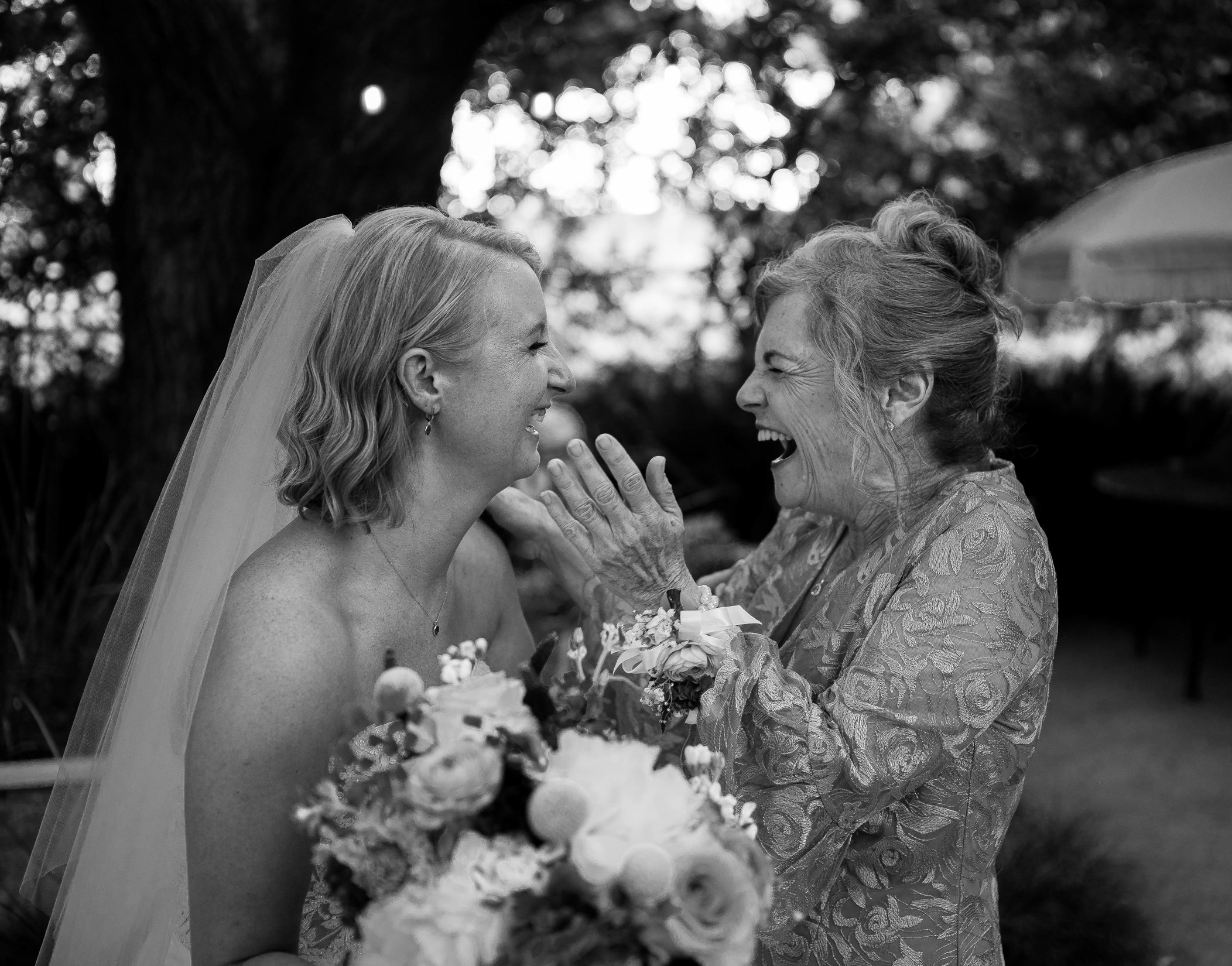 A bride and her mother share a joyful moment on a wedding day in an outdoor setting with trees in the background. 