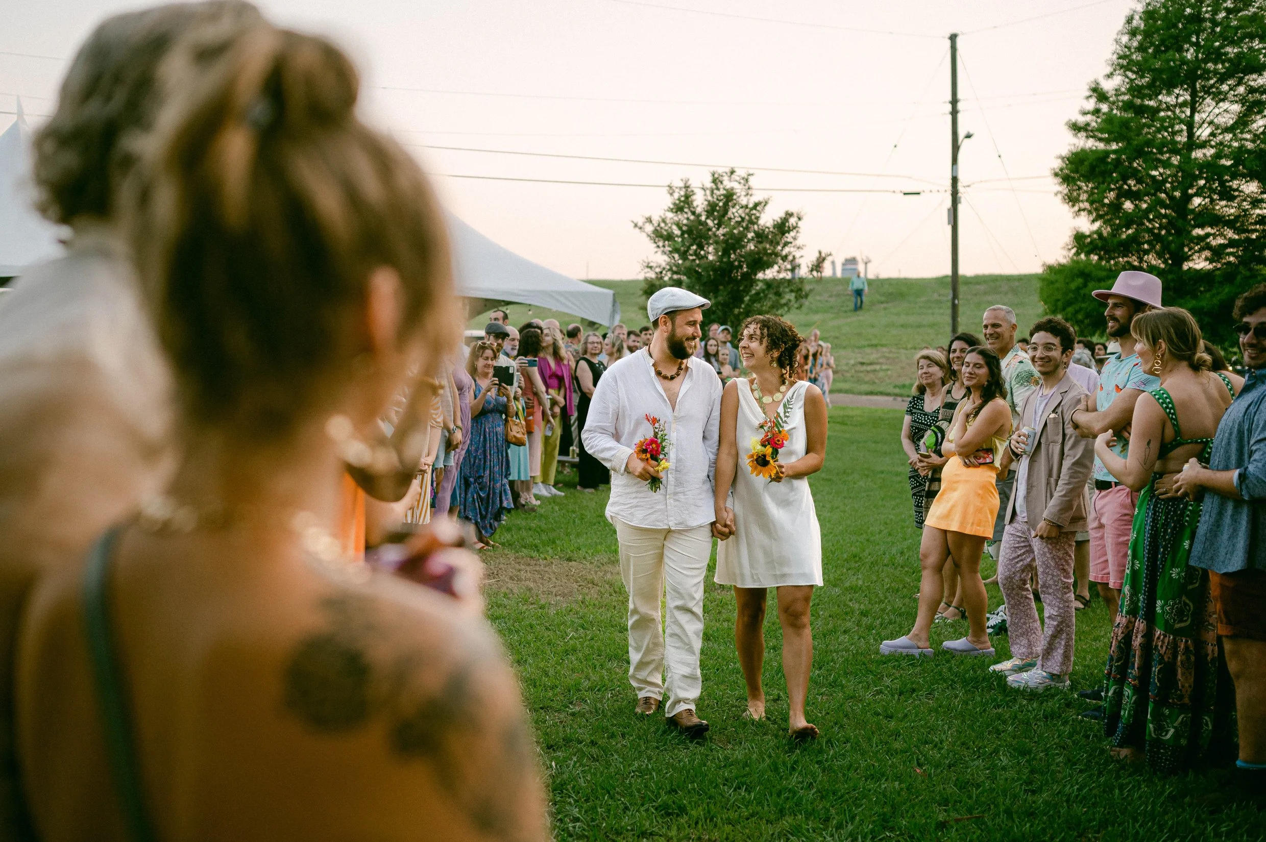 A couple holding hands and smiling at each other at an outdoor wedding ceremony with a crowd of guest watching, on a grassy area with trees and power lines in the background.