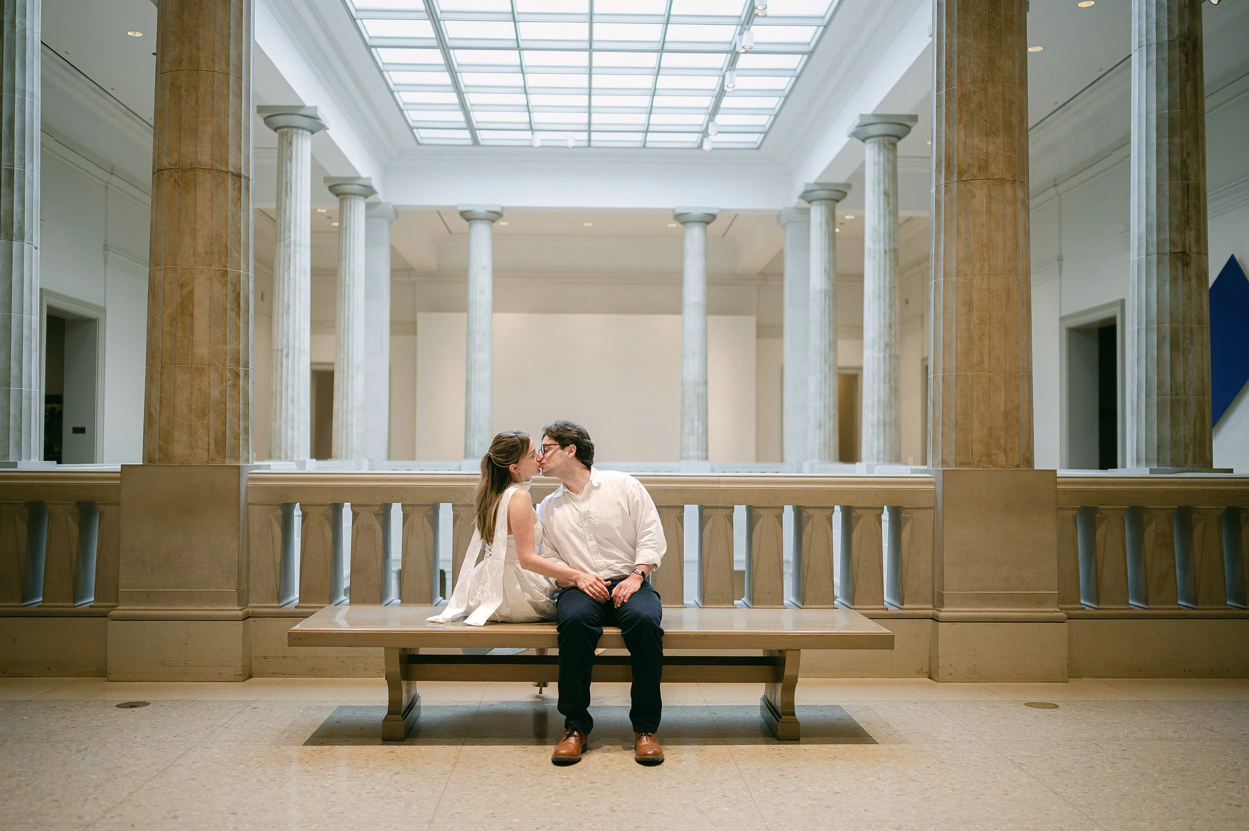 An engaged couple sitting on a wooden bench in the Art Institute of New Orleans with tall marble columns, a glass ceiling, and white walls.