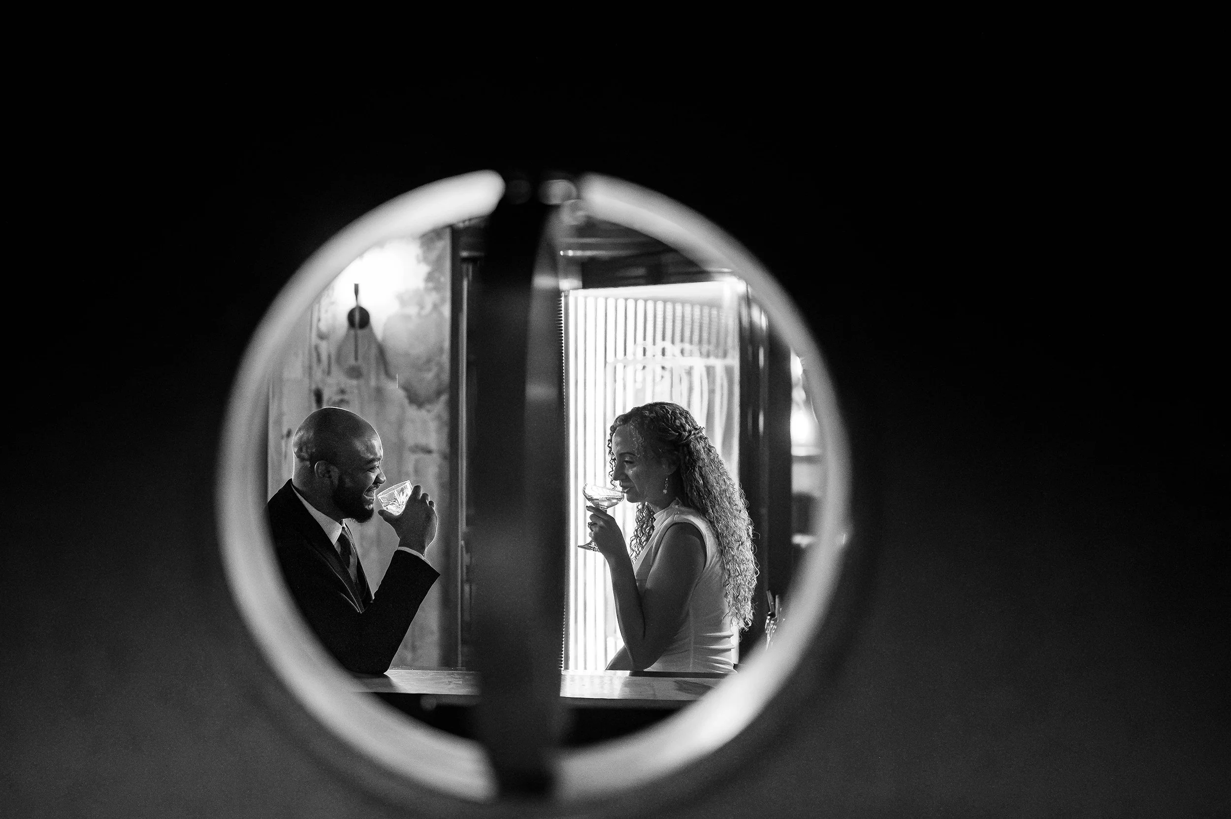 A black and white photo of an engaged couple through a circular window opening, both holding wine glasses and smiling.