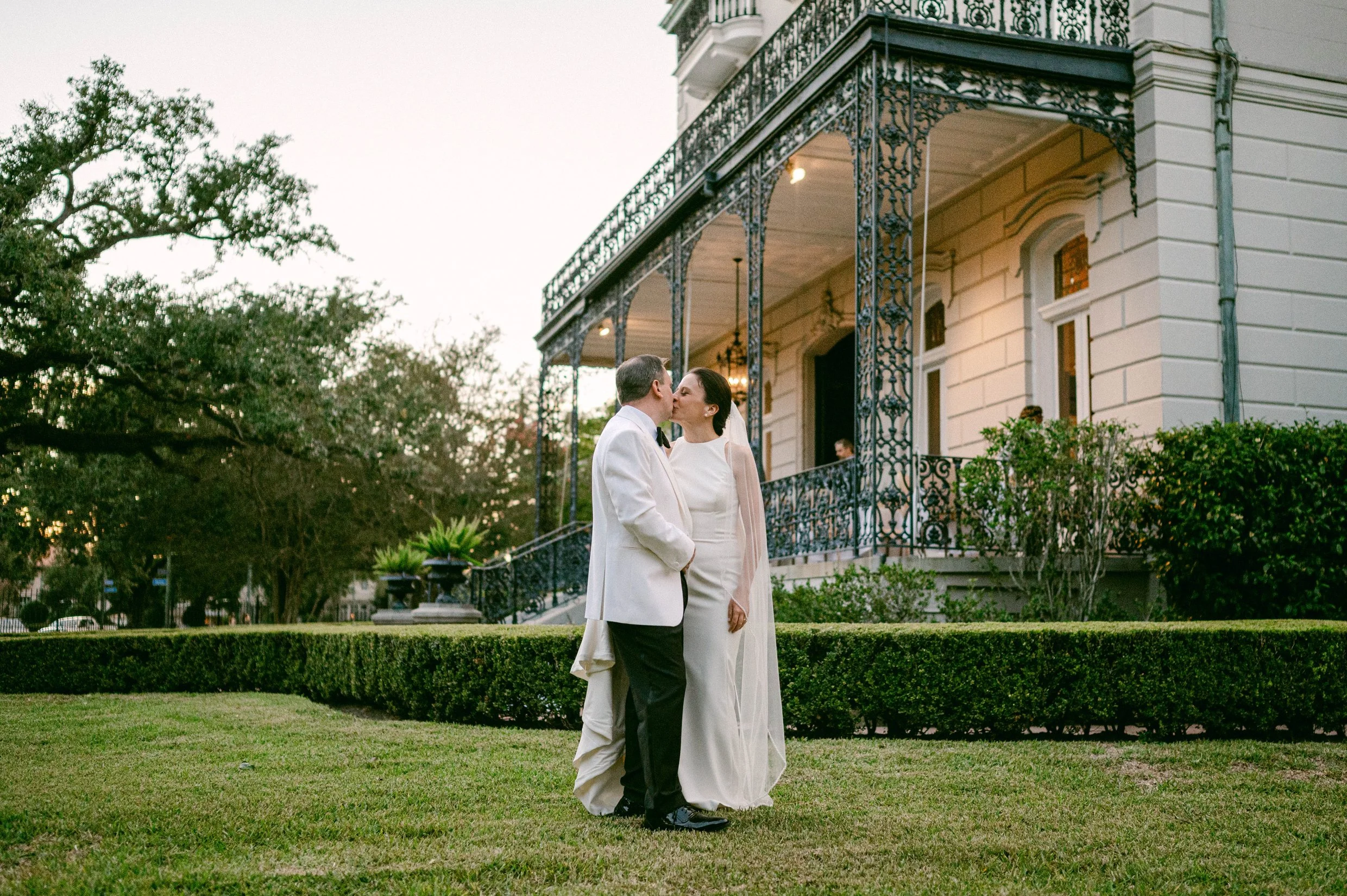 A couple dressed in wedding attire sharing a kiss outdoors in front of a historic New Orleans home with ornate black iron balconies and lush greenery.