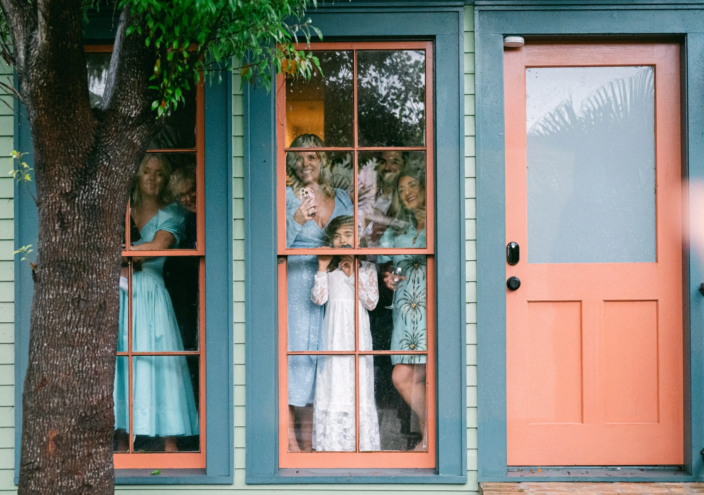 The bride and groom's families smiling and looking out through a house window, with a tree in the foreground.