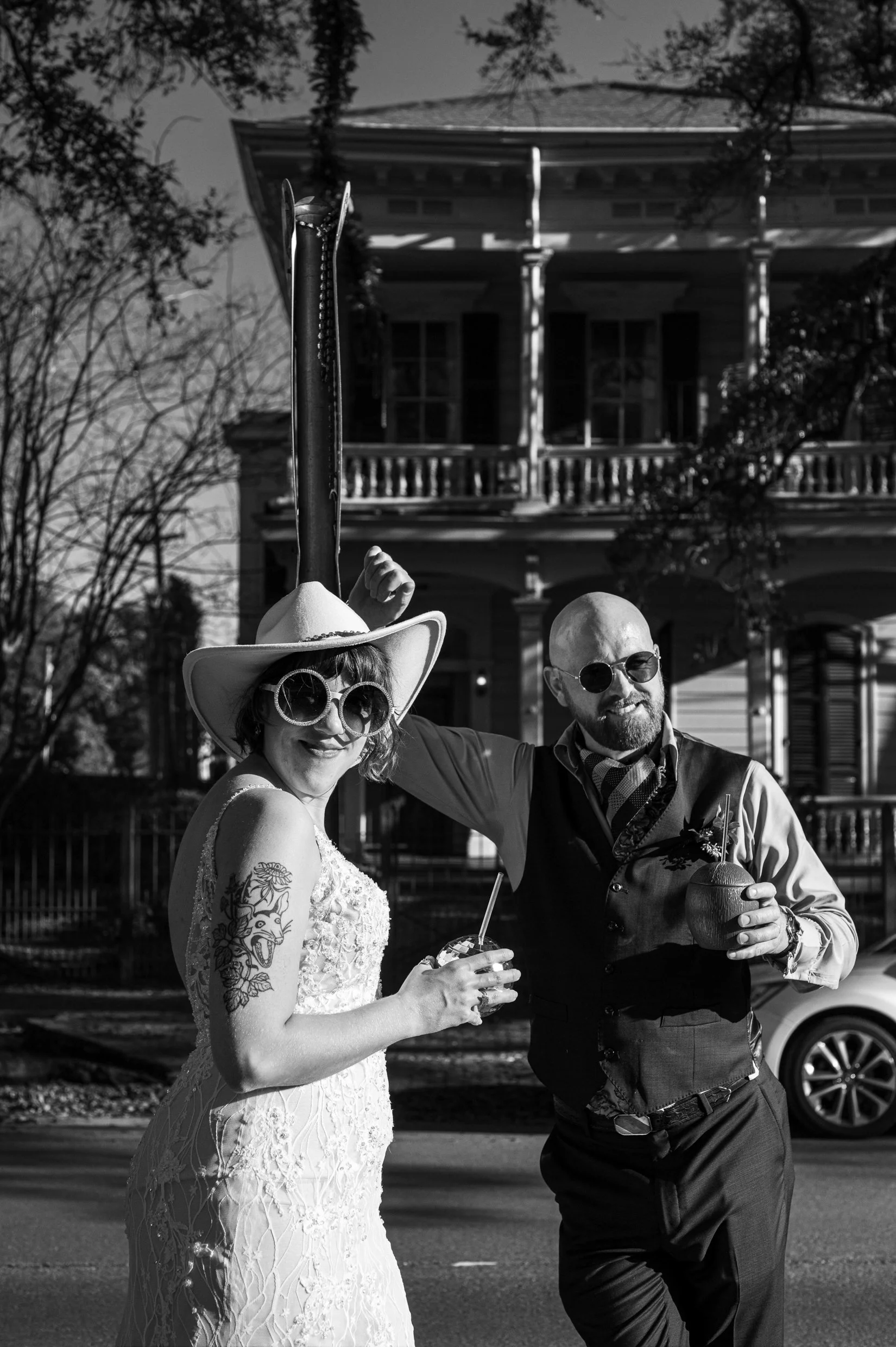 A black and white photo of a bride in a lace wedding dress and cowboy hat and a groom in a vest, both smiling and holding drinks.