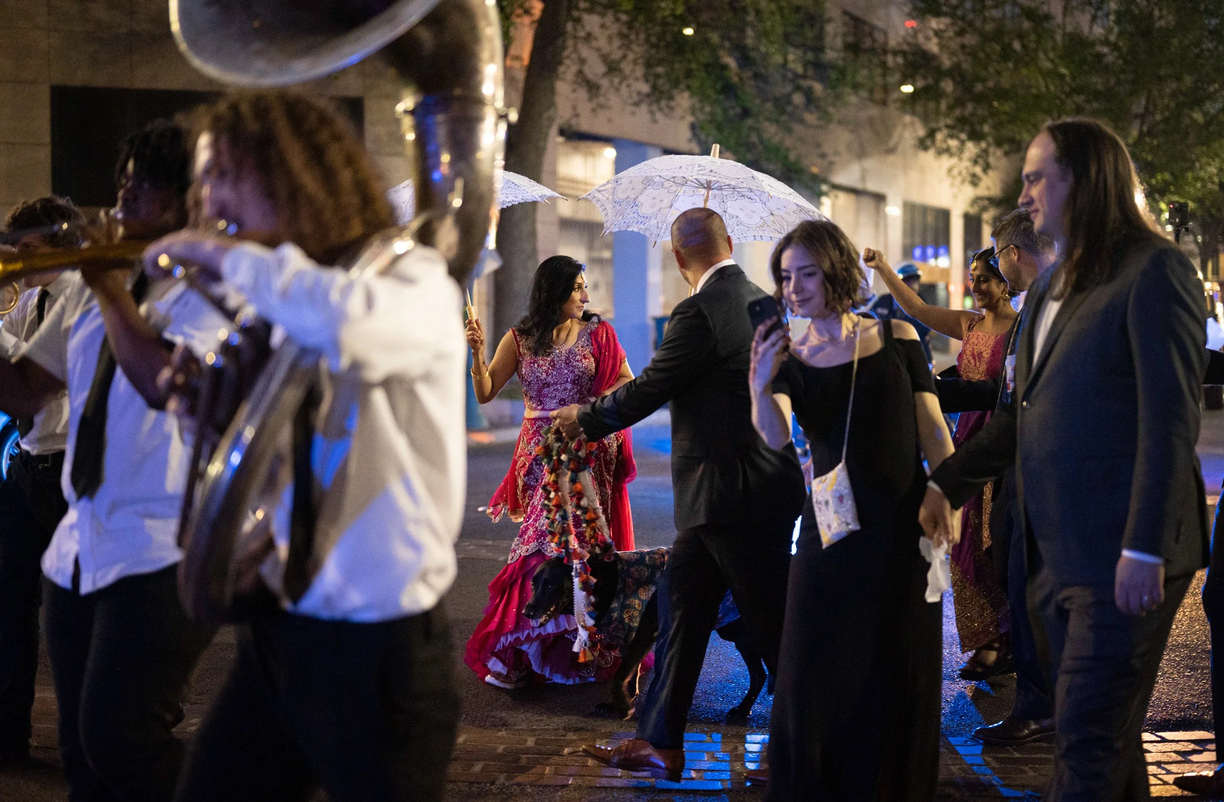 People in a New Orleans second line in traditional Indian wedding clothes, at night with umbrellas and trees in the background.