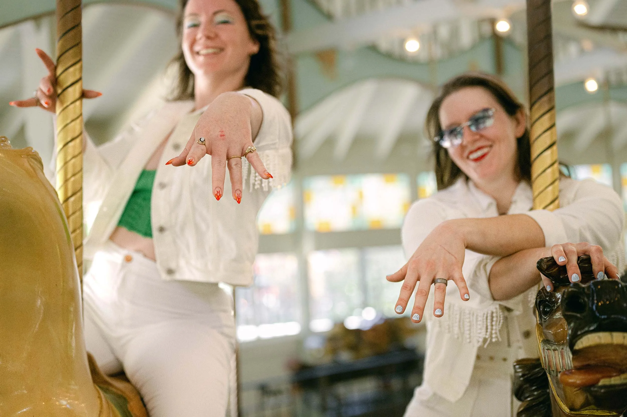 Two newlywed women at an amusement park carousel, showing off their wedding rings to the camera. 