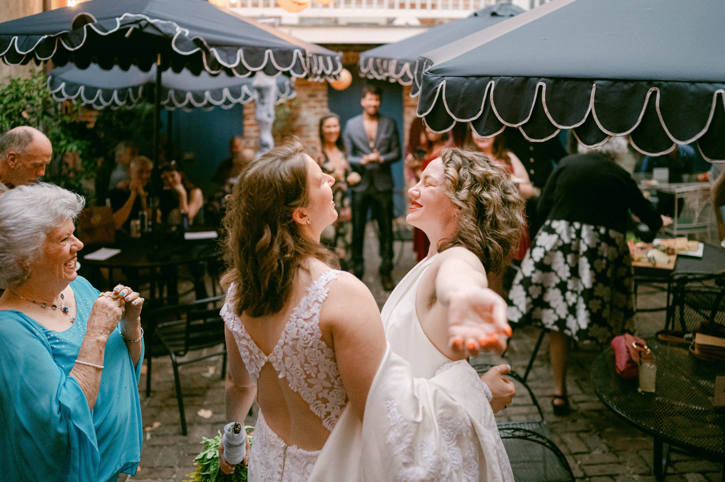 Two women, one in a lace wedding dress and the other in a white dress, dancing at their wedding, surrounded by guests under dark patio umbrellas in an outdoor setting.
