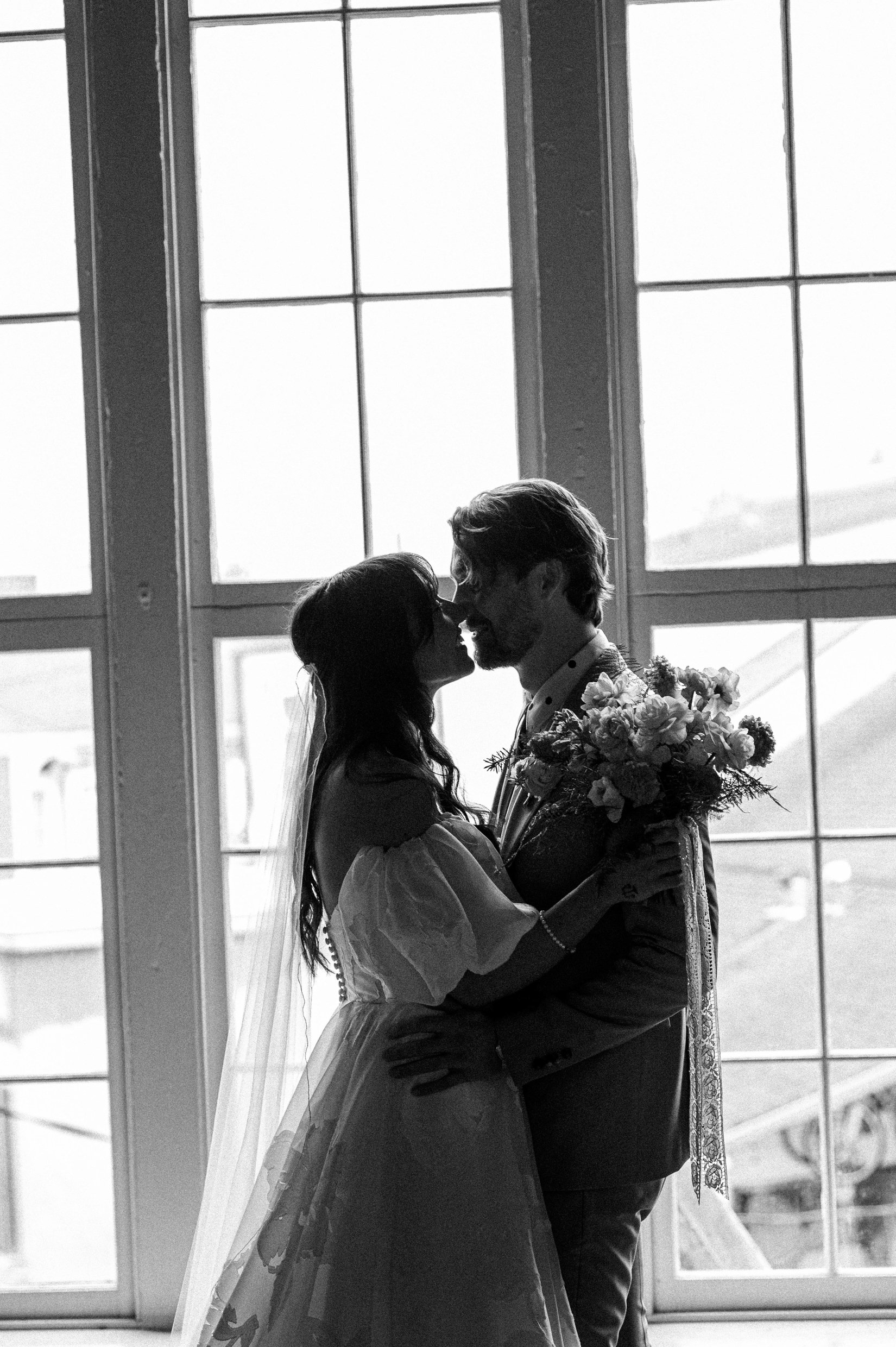 Silhouette of a bride and groom sharing a kiss indoors near large windows.