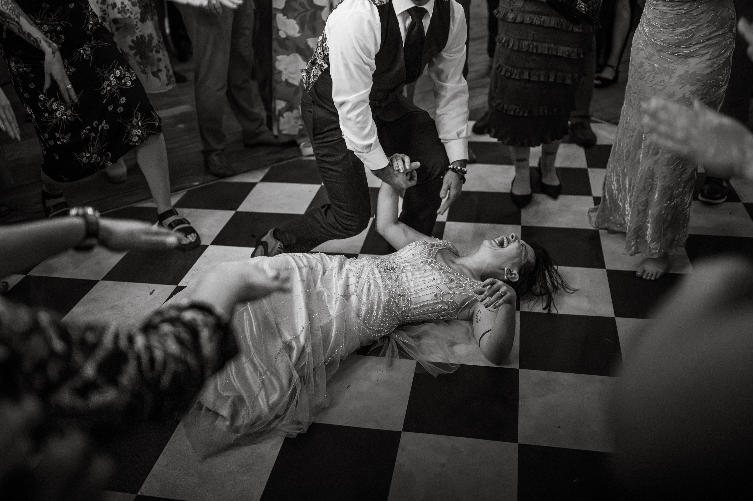 People dancing at a wedding reception, with a bride lying on a checkered floor and laughing and a groom holding her hand, surrounded by guests 