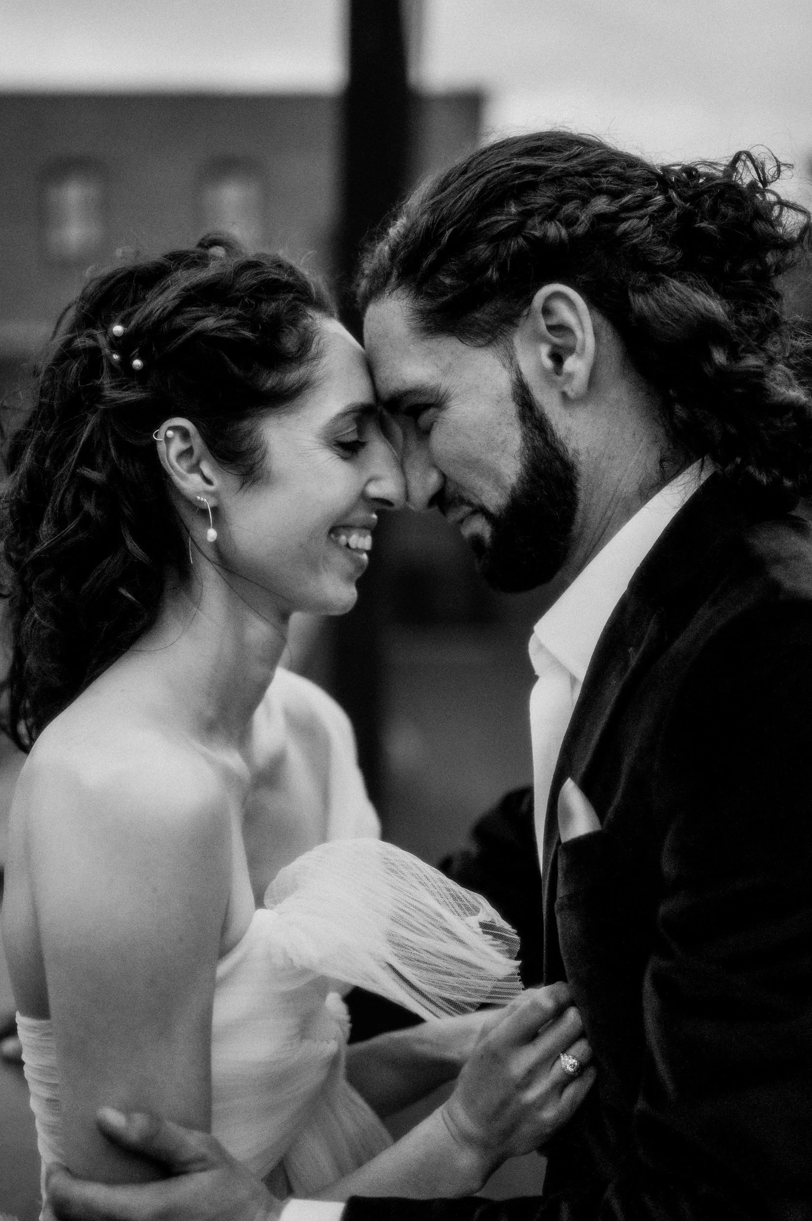 A black and white photo of newlyweds sharing a close, intimate moment with their foreheads touching and smiling with eyes closed, dressed in wedding attire.