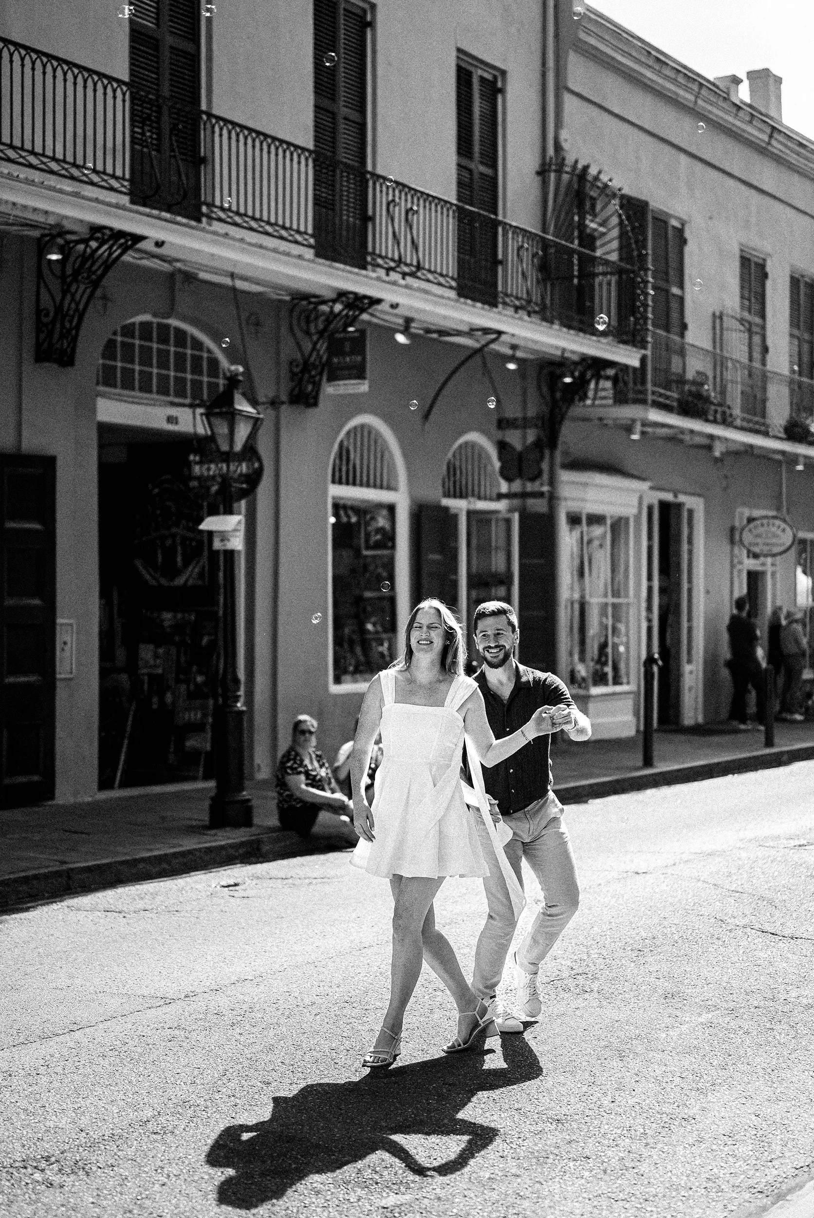 A couple walking hand in hand on New Orleans French Quarter street, smiling. There are store fronts and balconies with shutters behind them, and people sitting and walking nearby.