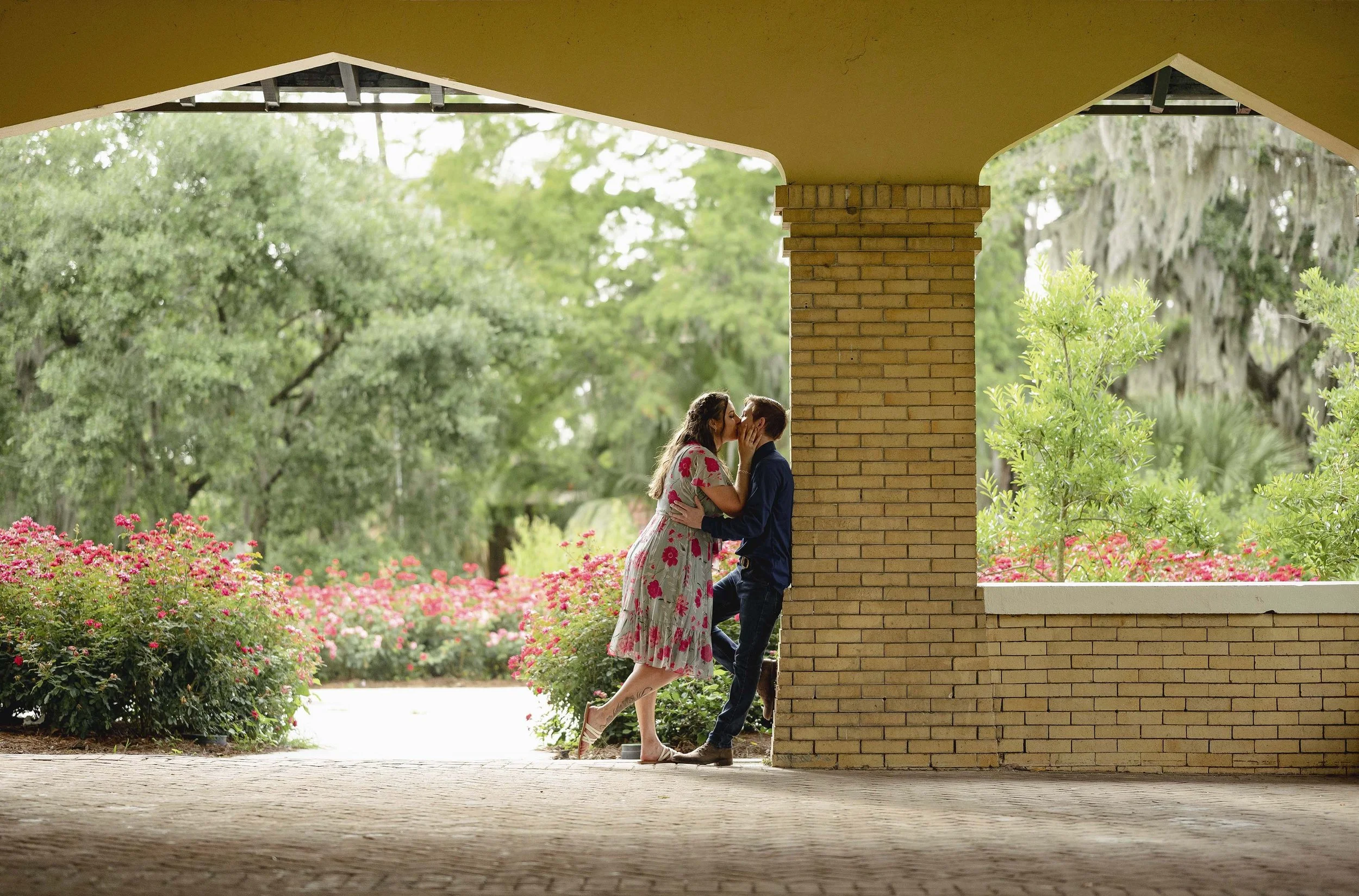 An engaged couple kissing under a covered New Orleans courtyard with brick columns, surrounded by blooming pink flowers and green trees in the background.
