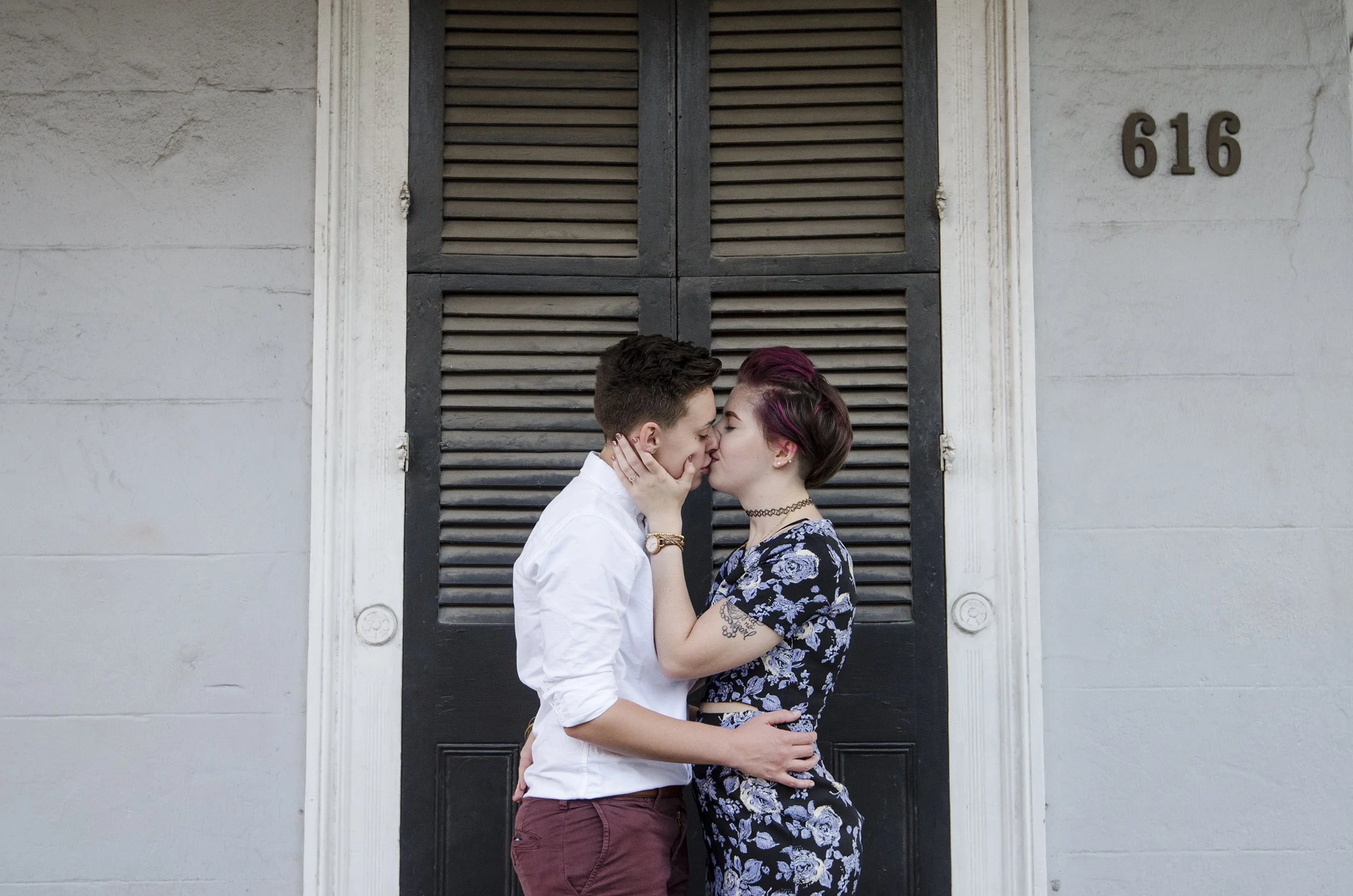 A young queer couple kissing in front of a black shuttered window on a light gray wall in New Orleans French Quarter.