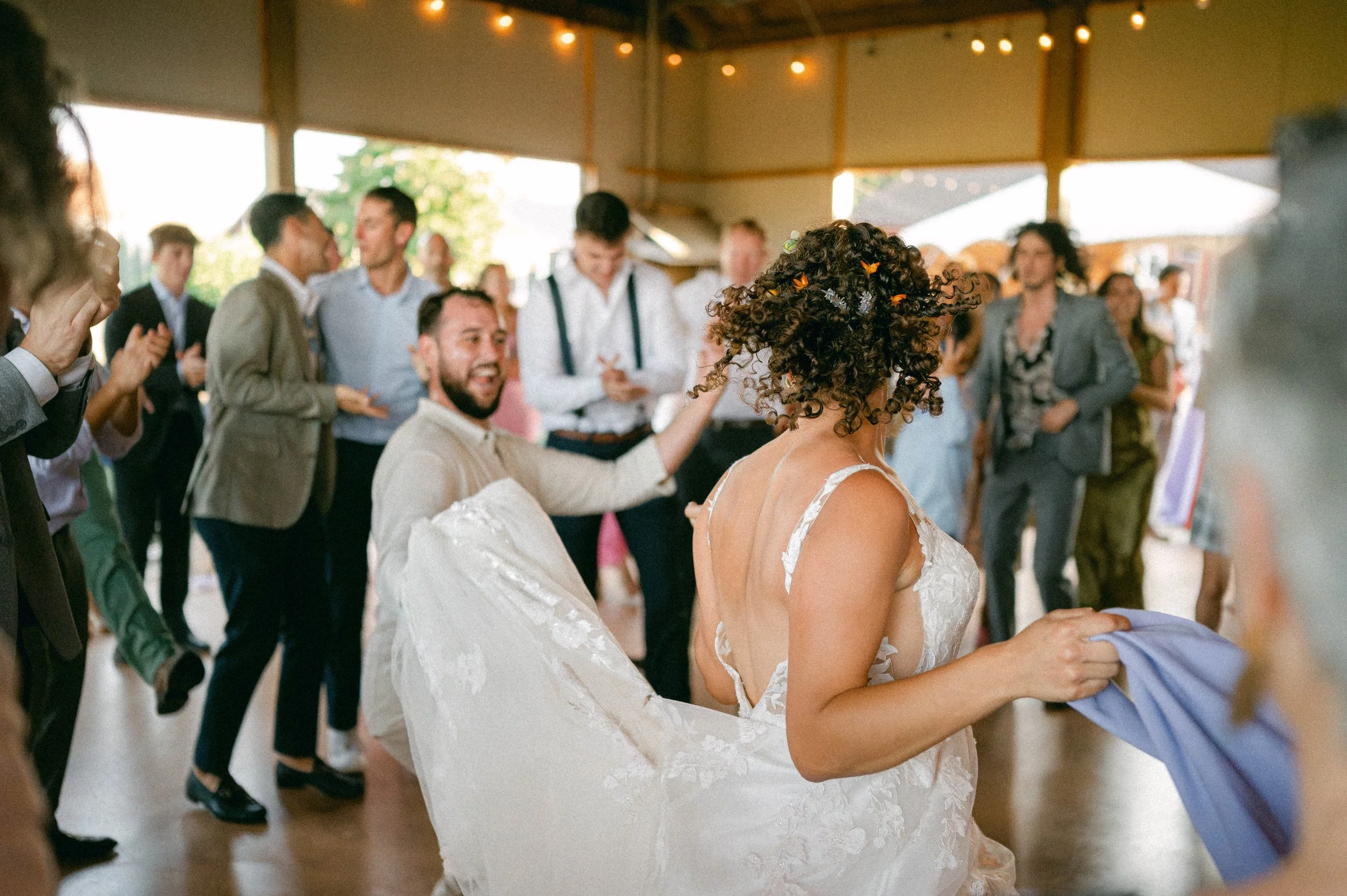 People dancing and celebrating at a wedding reception, with a bride in a white lace wedding dress and a groom in a beige suit.