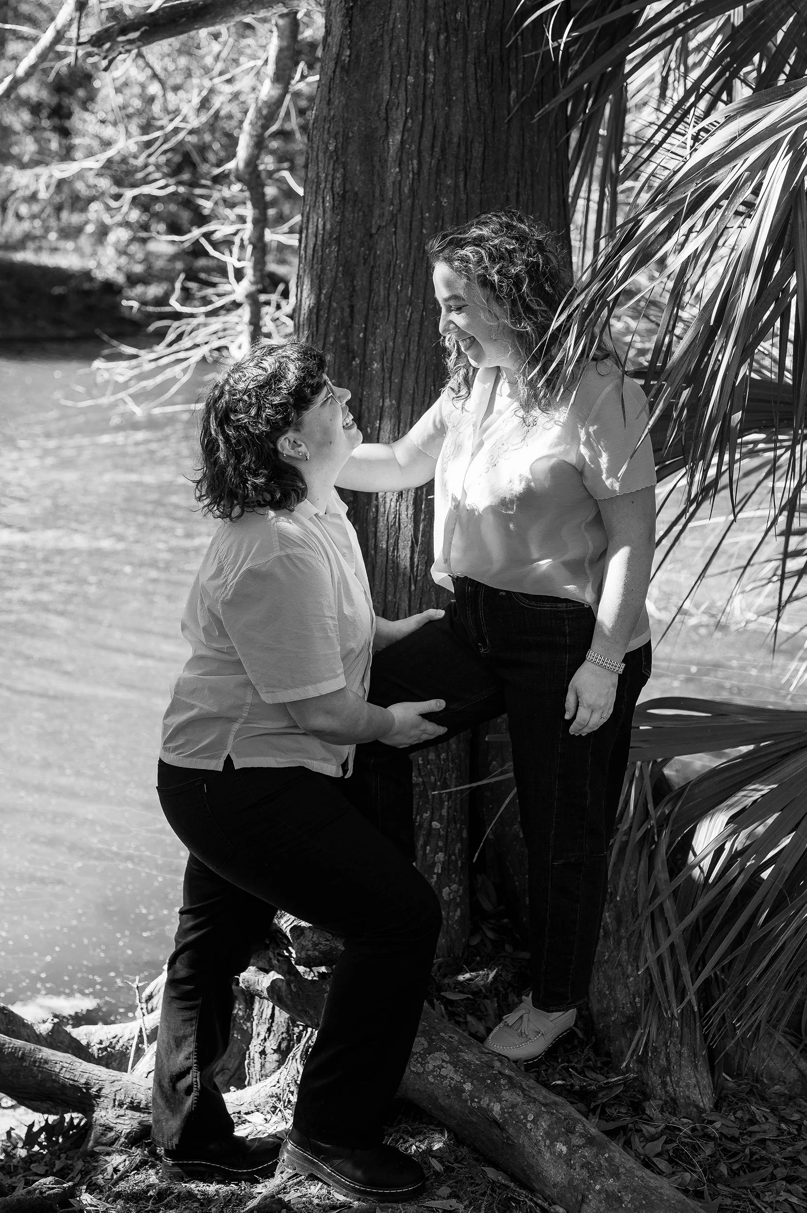 Two women smiling and looking at each other, standing outdoors by a tree near a body of water, in a black-and-white photo.