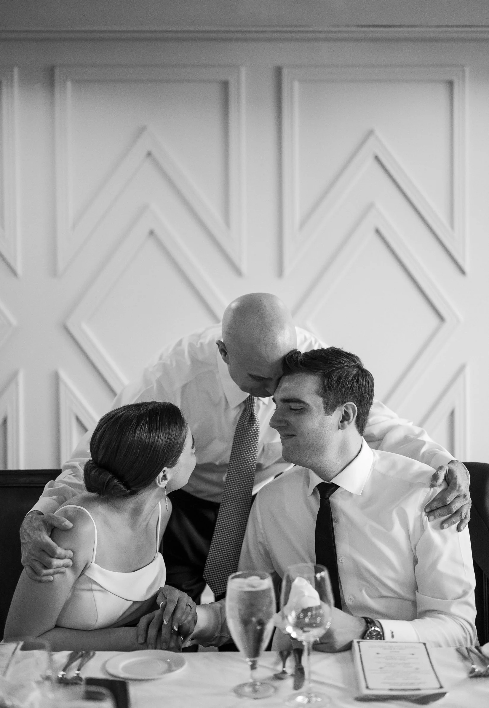 A pair of newlyweds and the groom's father are gathered closely at a formal dinner table. The father is kissing the groom on the forehead, and the bride is looking at them with a smile. 