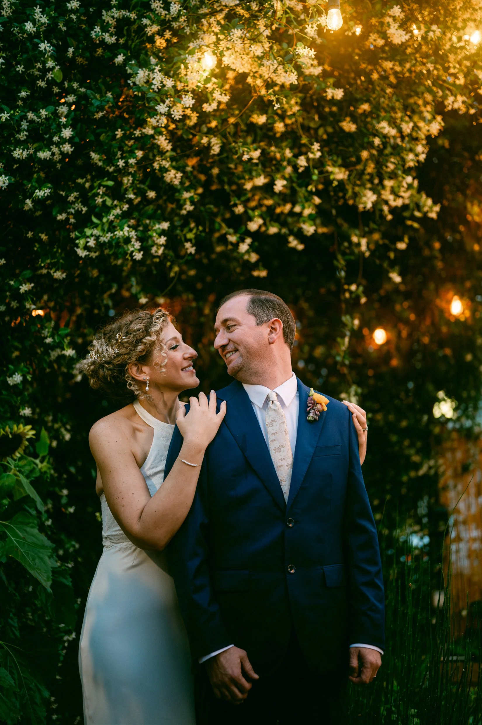 A bride and groom smiling and looking at each other outdoors during sunset, surrounded by greenery and illuminated by string lights and warm lighting.