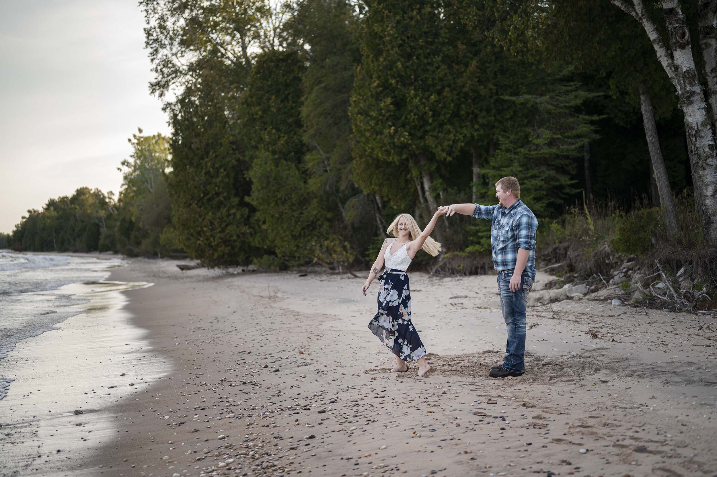 A young couple dancing on a sandy beach near Lake Michigan. They are holding hands and smiling, with large green trees and a cloudy sky in the background.