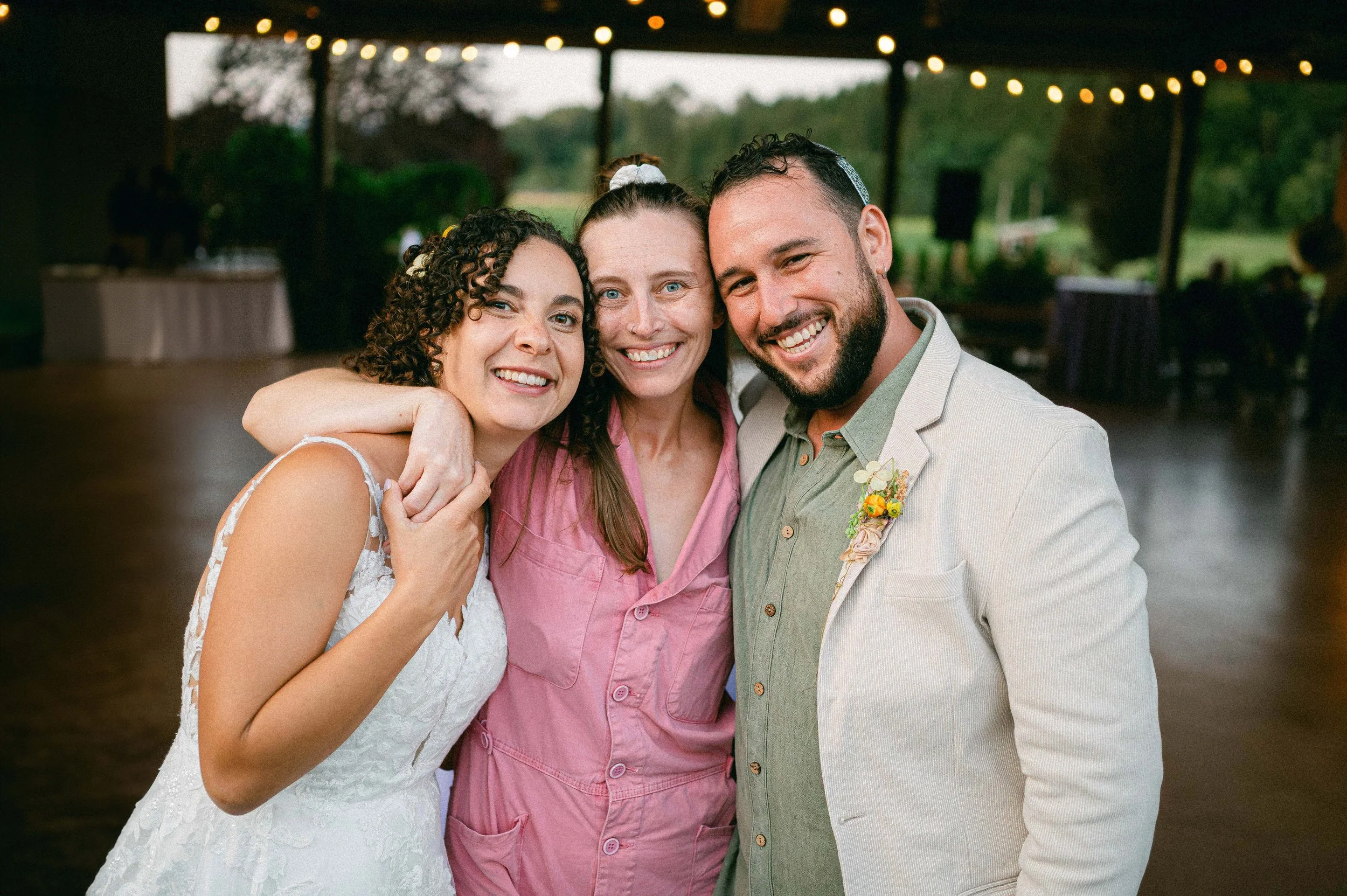 Wedding photographer stands next to bride and groom, hugging each other after wedding under string lights, with trees and a picnic table in the background.