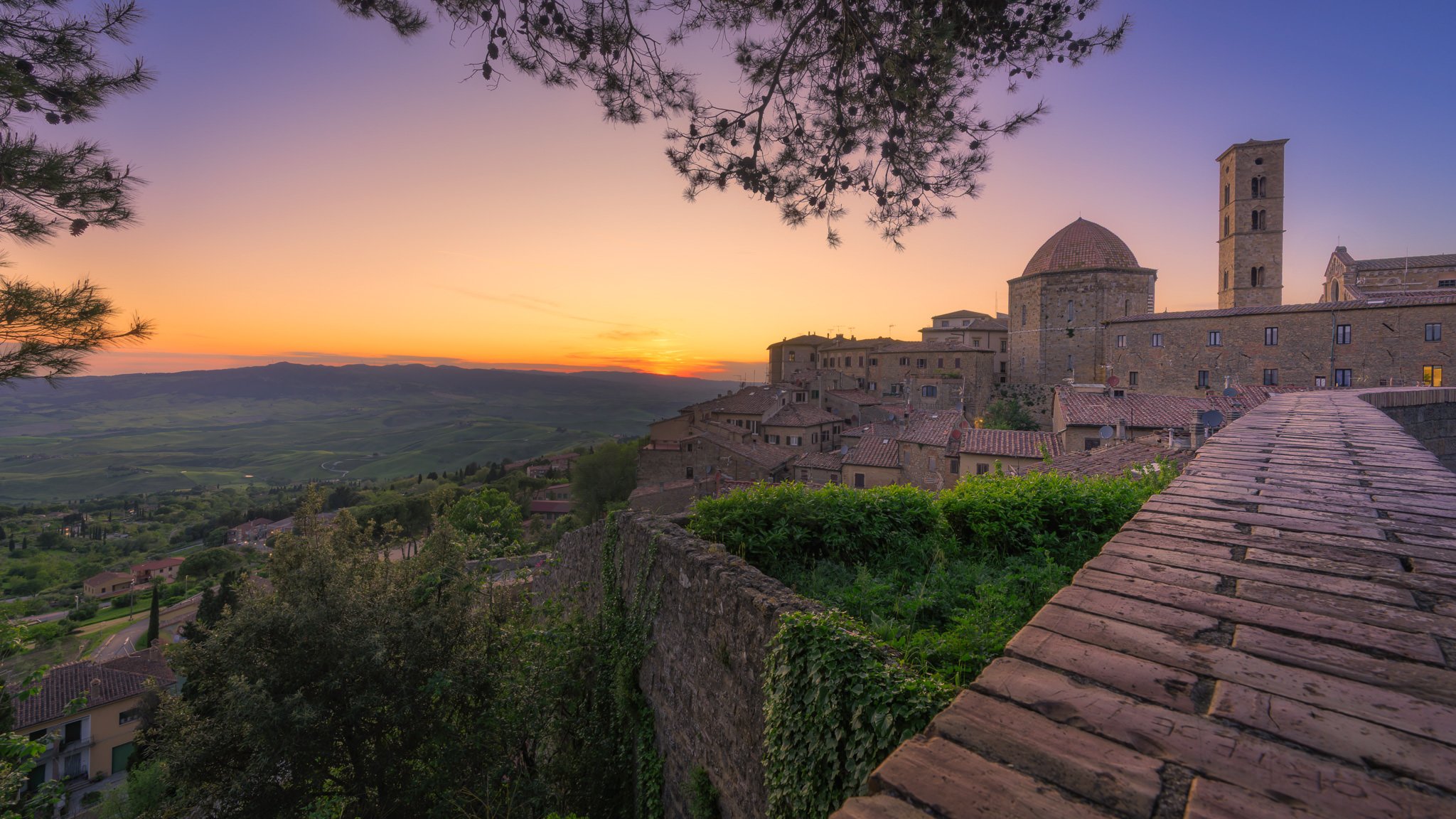 Sunset over a historic Italian village with stone buildings, tiled roofs, a dome, a tower, and a scenic landscape in the background.