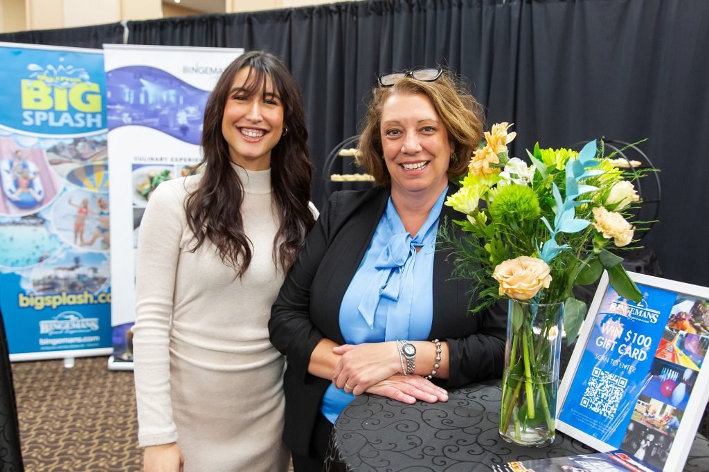 Two women smiling at an event, standing behind a table with a large floral arrangement in a glass vase and promotional materials for Big Splash water park.