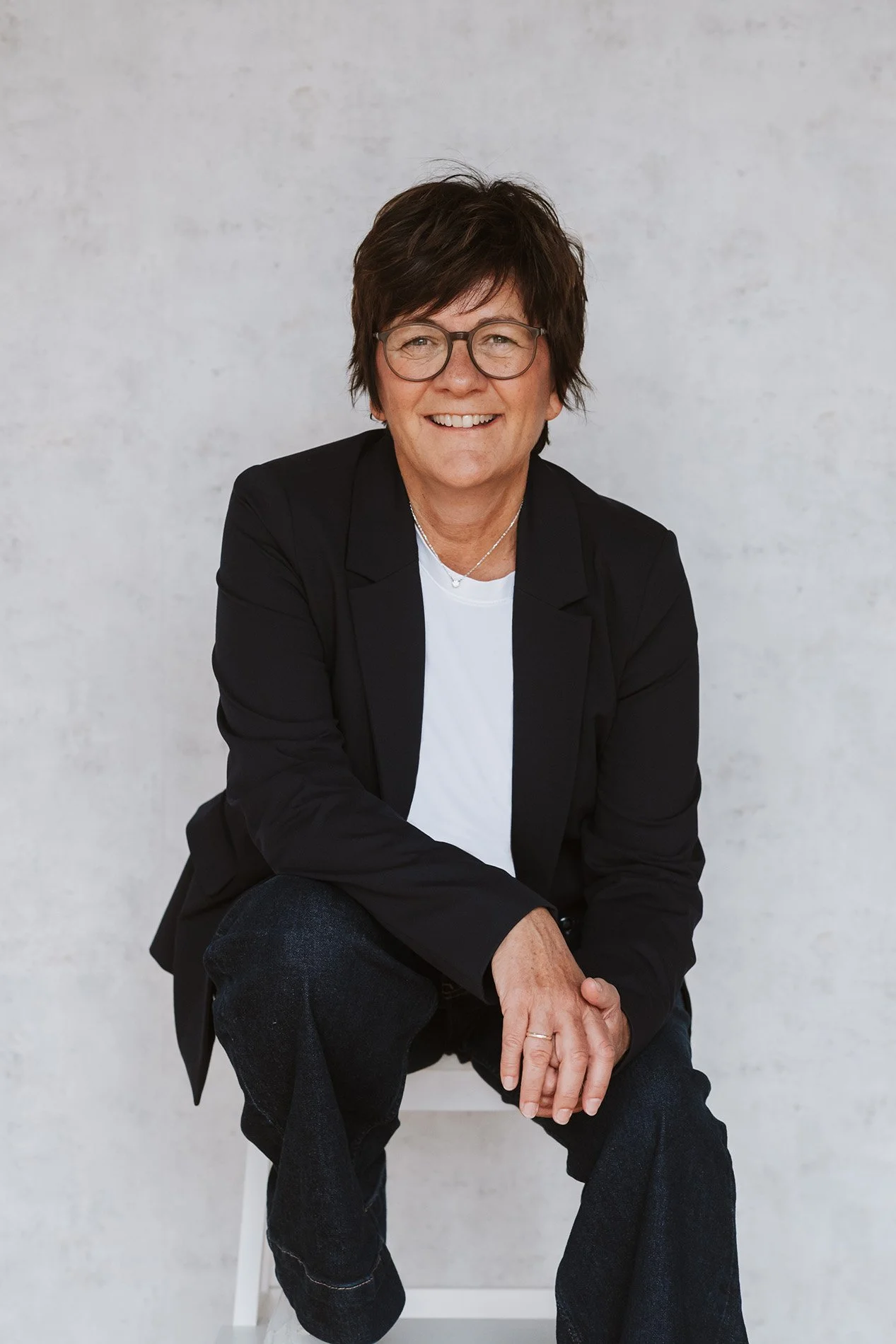 Onika mit Brille, dunkelbraunen Haaren, schwarzem Blazer, weißem T-Shirt und Jeans sitzt vor einem weißen Hintergrund.