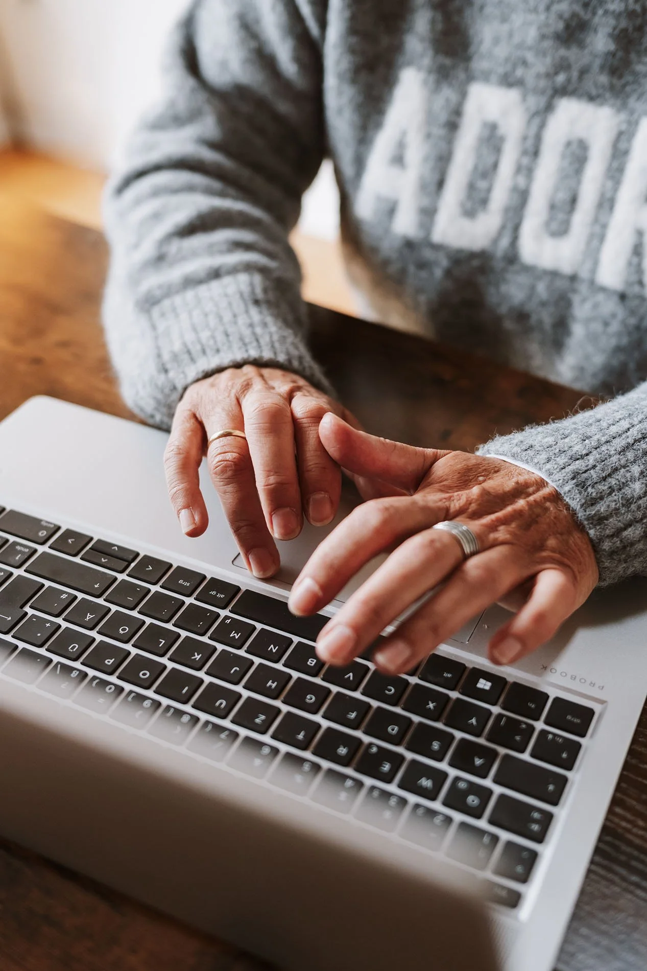 Onika tippt auf einem Laptop tippt, getragen von einem grauen Pullover, sitzend an einem Holztisch.
