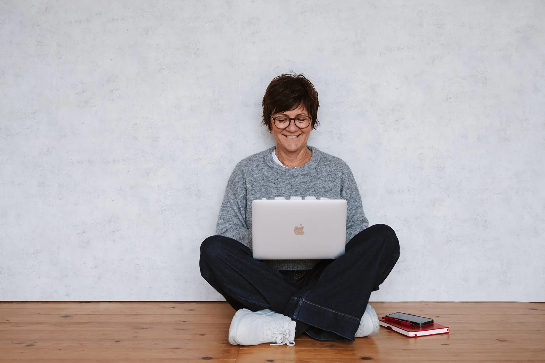 Onika sitzt auf dem Boden vor einer weißen Wand, schaut lachend auf einen offenen Laptop auf ihrem Schoß, mit Notizbuch und Smartphone neben ihr.