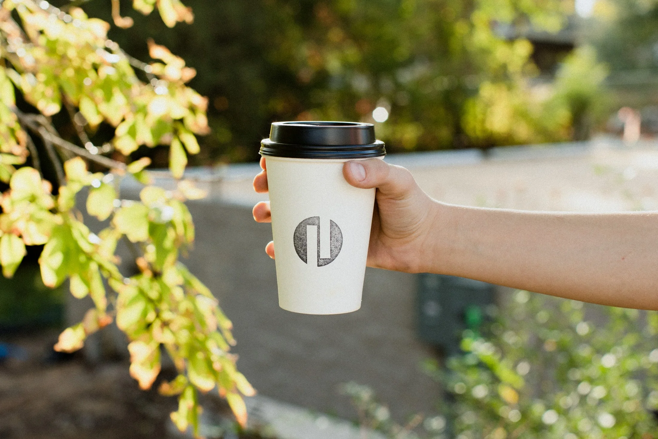 White Coffee cup with black logo. Held in front of greenery