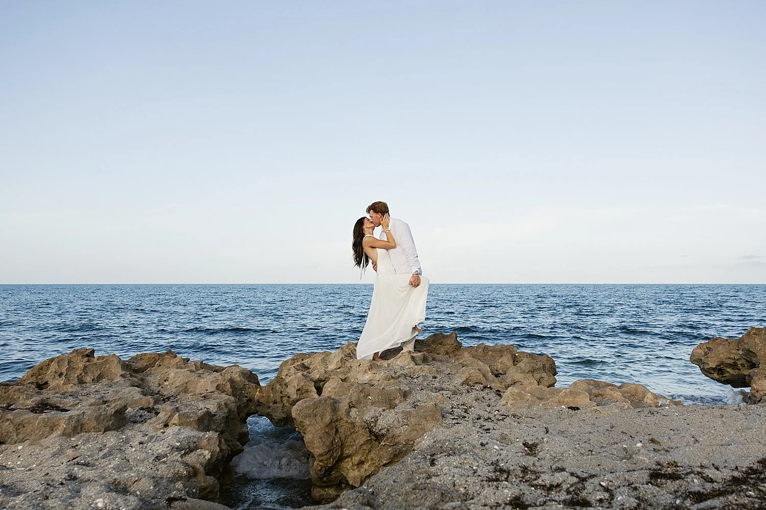  groom-to-be leans in to kiss his bride on ocean rocks by Florida wedding photographer 