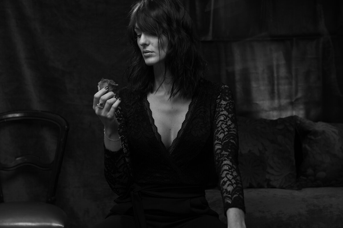 Laurie Codaccioni, plongée dans ses pensées, tient une rose fanée dans un décor dramatique. Le jeu de textures entre la dentelle de sa tenue et le décor de fond ajoute de la profondeur à l'image, accentuée par l'éclairage doux et contrasté qui met e