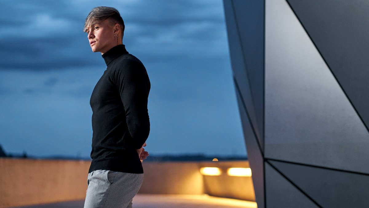  Dans cette image élégante et contemporaine, Tristan Defeuillet Vang, YouTuber et influenceur, pose sur les terrasses modernes du musée Confluence à Lyon. Il est vêtu d’un col roulé noir ajusté et d’un pantalon clair à carreaux, dégageant une allure 