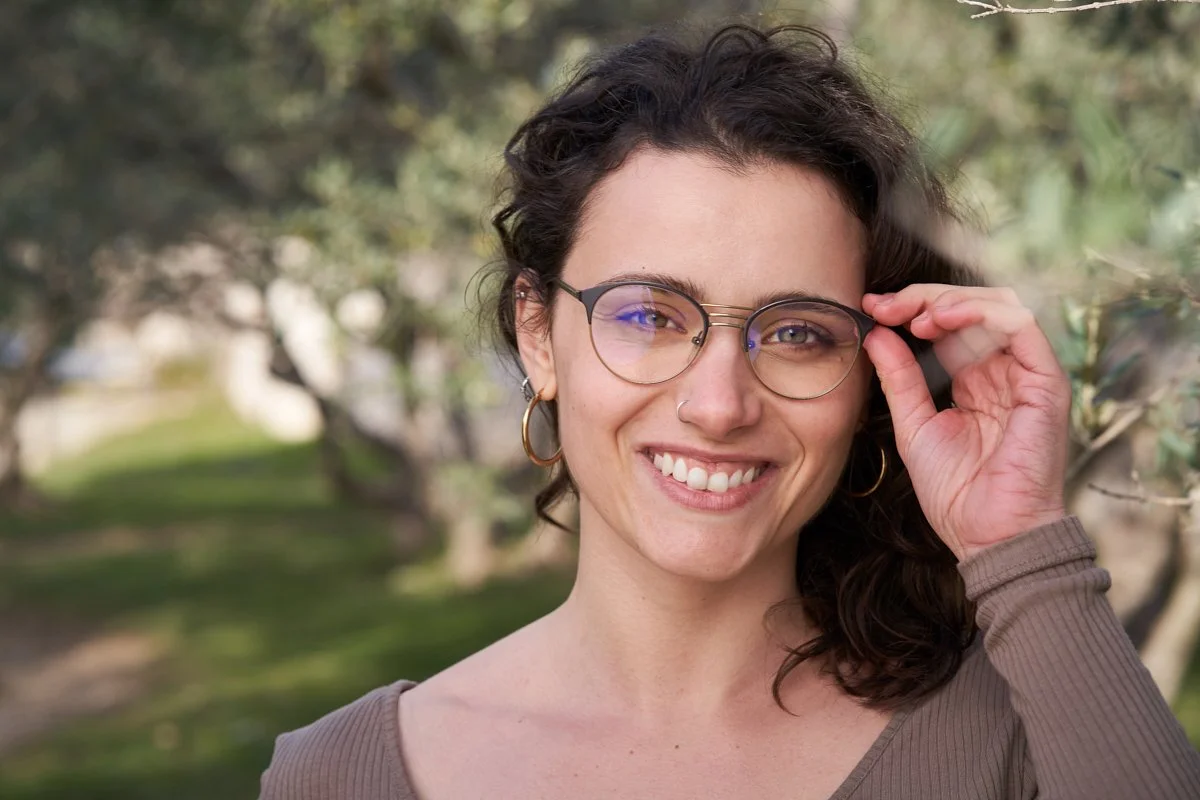  Ce portrait de Gersende Rambaud, réalisé en extérieur, met en valeur son sourire chaleureux et naturel. Elle porte des lunettes au design moderne, qu'elle ajuste légèrement avec sa main. La lumière naturelle illumine son visage et accentue l’éclat d