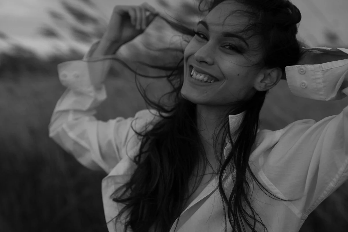  Ce portrait en noir et blanc de Catherine Scaramuzzi capture un moment de spontanéité et de joie naturelle. La modèle, habillée d'une chemise blanche légèrement déboutonnée, rit avec une expression lumineuse, ses mains levées vers ses cheveux, pris 