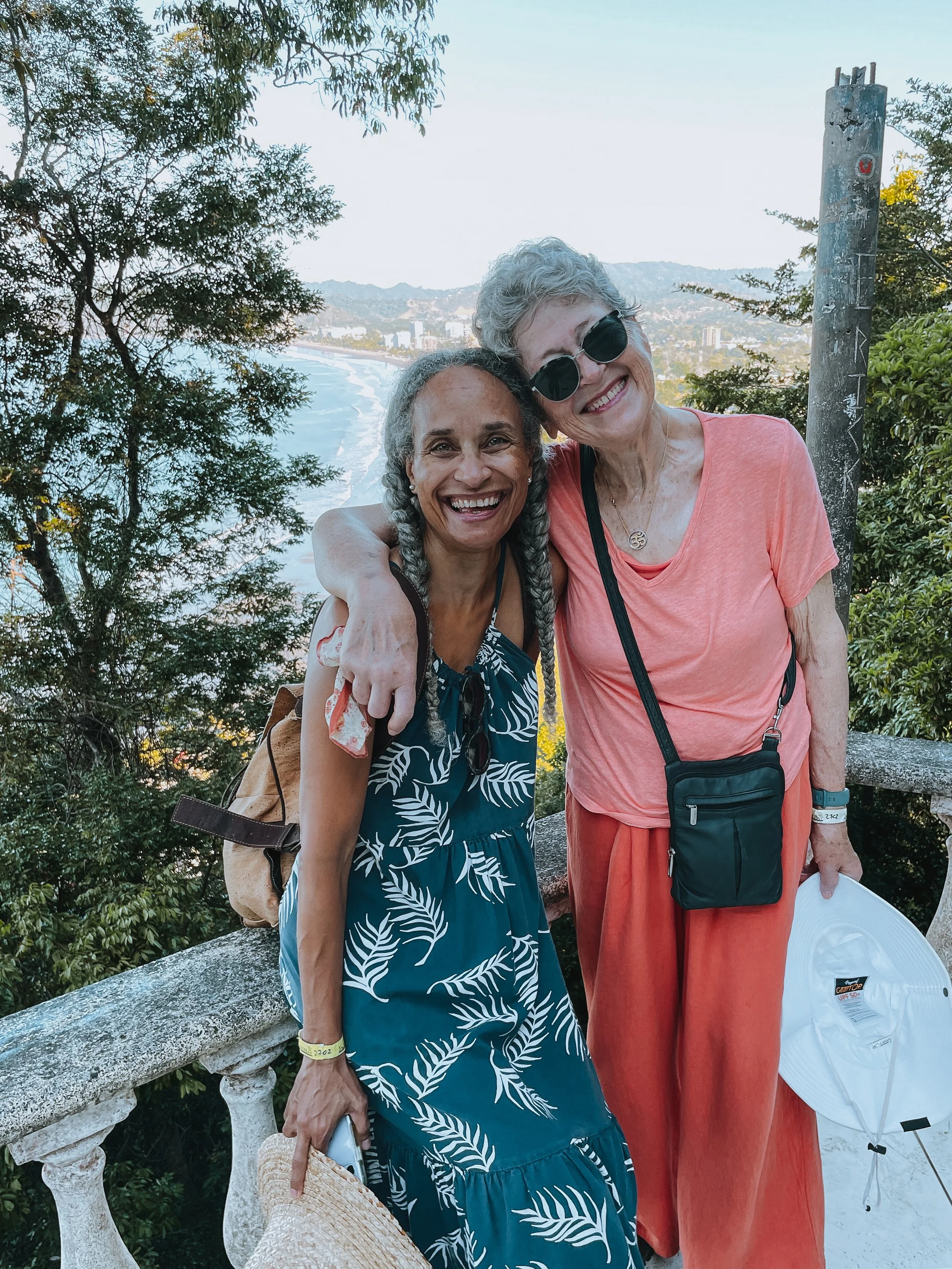 Two women smile and pose in front of a scenic ocean view, surrounded by trees and greenery, with a distant city and hills in the background.