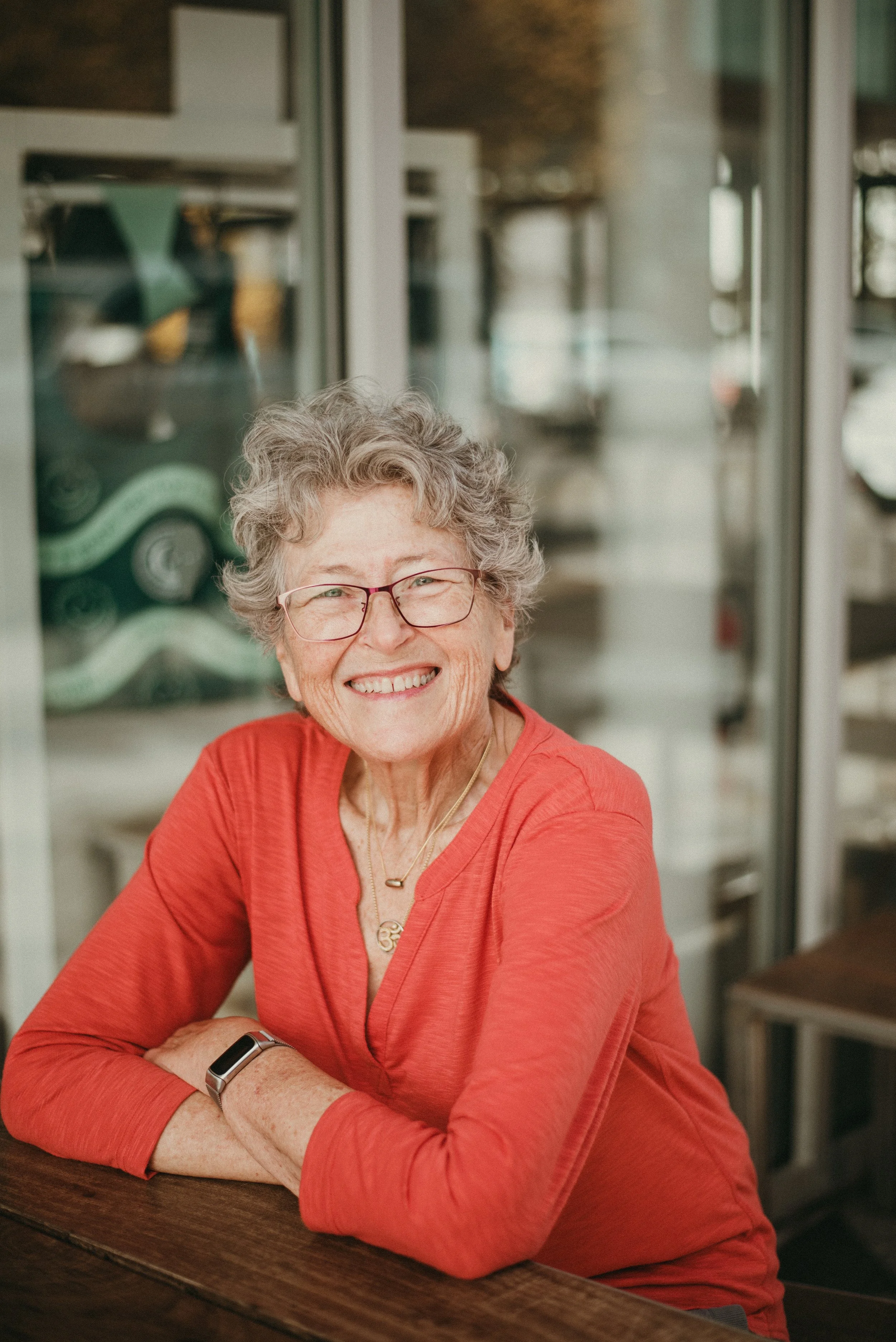 Smiling elderly woman with glasses, curly gray hair, and a red shirt sitting at a wooden table in a cozy cafe or restaurant.