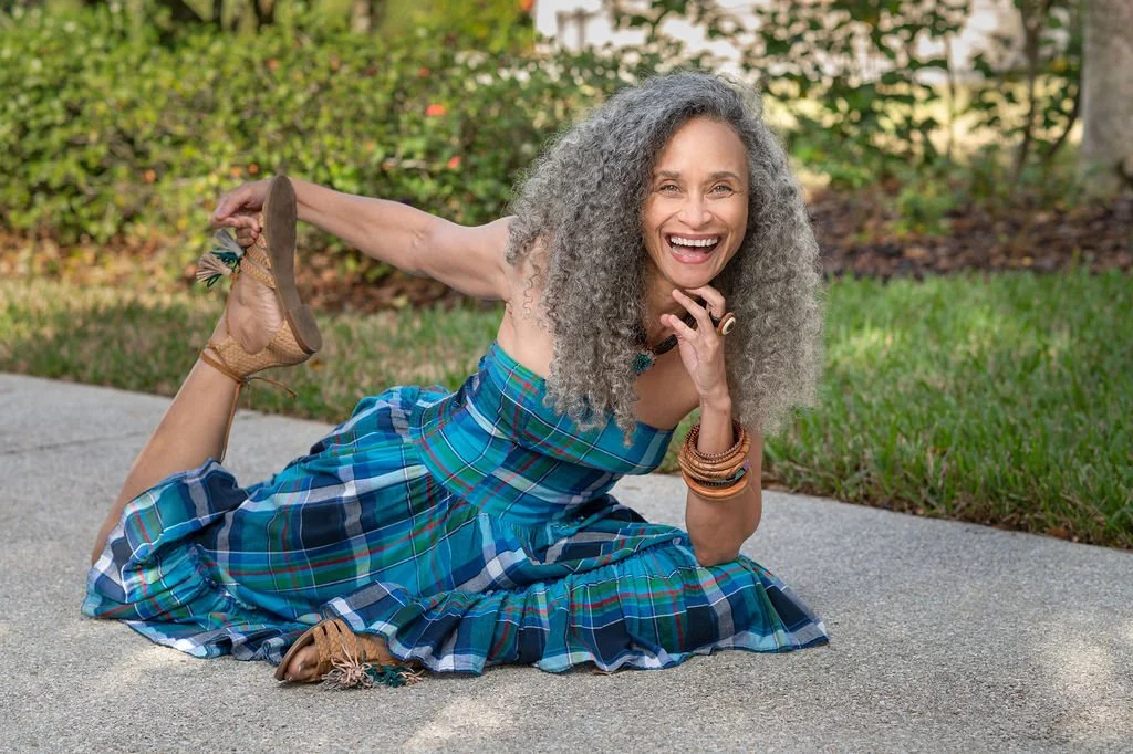 Smiling woman with gray curly hair lying on the sidewalk outdoors, wearing a blue plaid dress and holding a sandal behind her back.