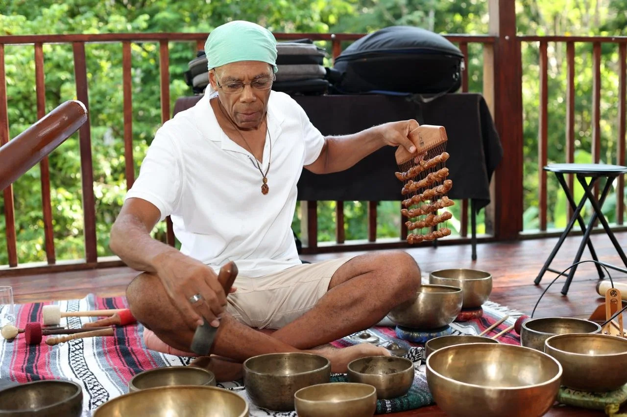 An elderly man sitting cross-legged on a rug on a wooden deck, playing steel drums and holding a wooden percussion instrument, with singing bowls and other instruments around him, surrounded by green trees.
