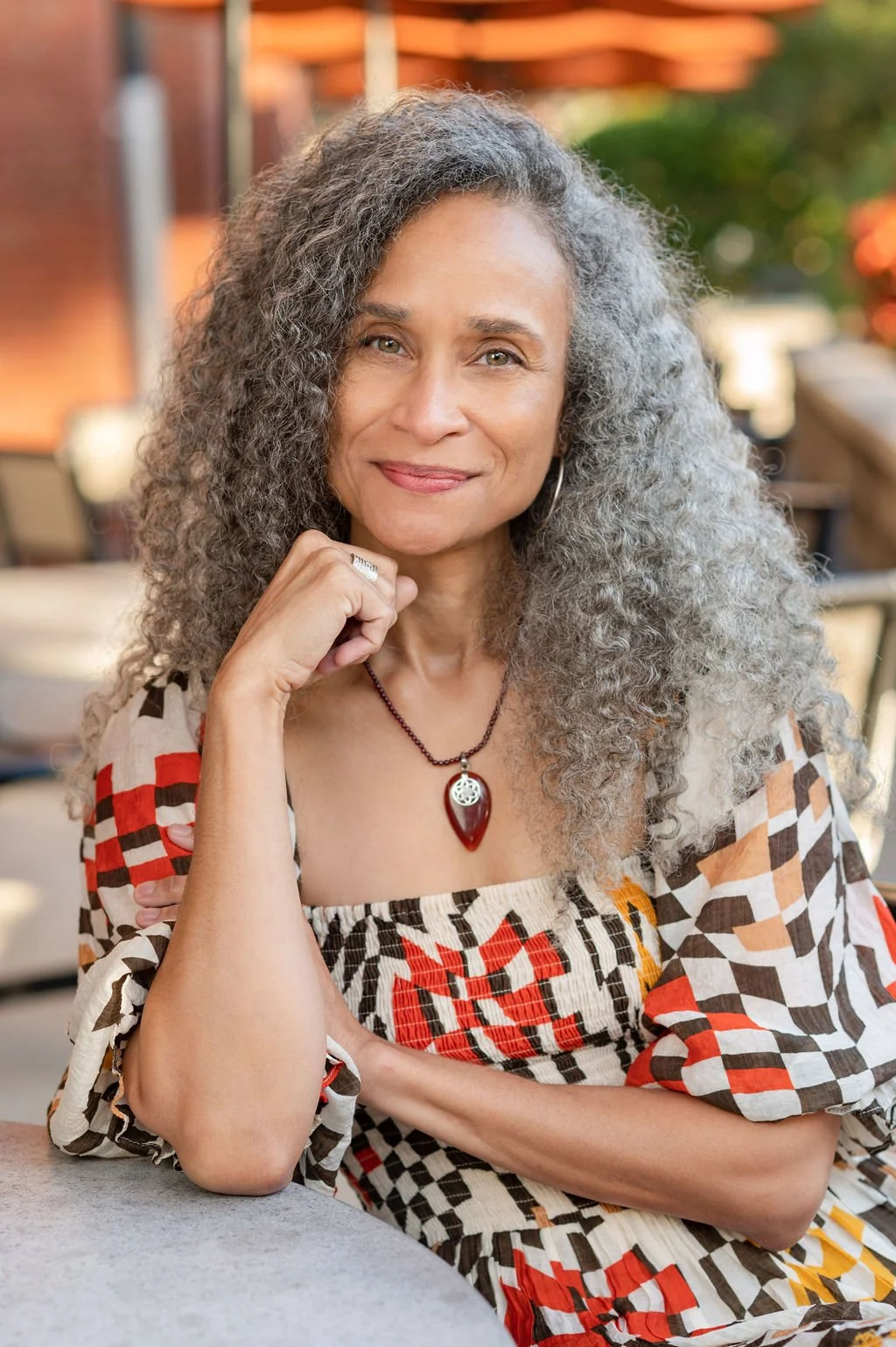 A woman with curly gray hair sitting outdoors at a table, wearing a patterned dress and a necklace with a large red teardrop pendant, smiling gently.